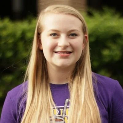 Young girl with long blonde hair wearing a purple shirt, smiling outdoors with greenery in the background.