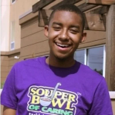Young man smiling outdoors wearing a purple T-shirt with a Souper Bowl of Caring logo.