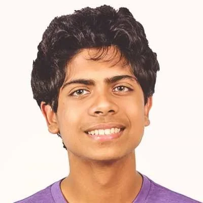A young man with curly black hair smiling, wearing a purple shirt against a plain light background.