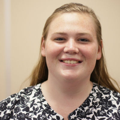 A young woman with long, light brown hair and a cheerful smile, wearing a black and white patterned shirt against a beige background.