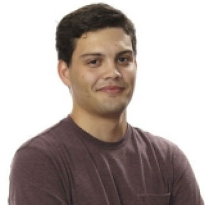 A young man with short dark hair wearing a burgundy shirt, smiling with one arm crossed over his chest, facing the camera against a white background.