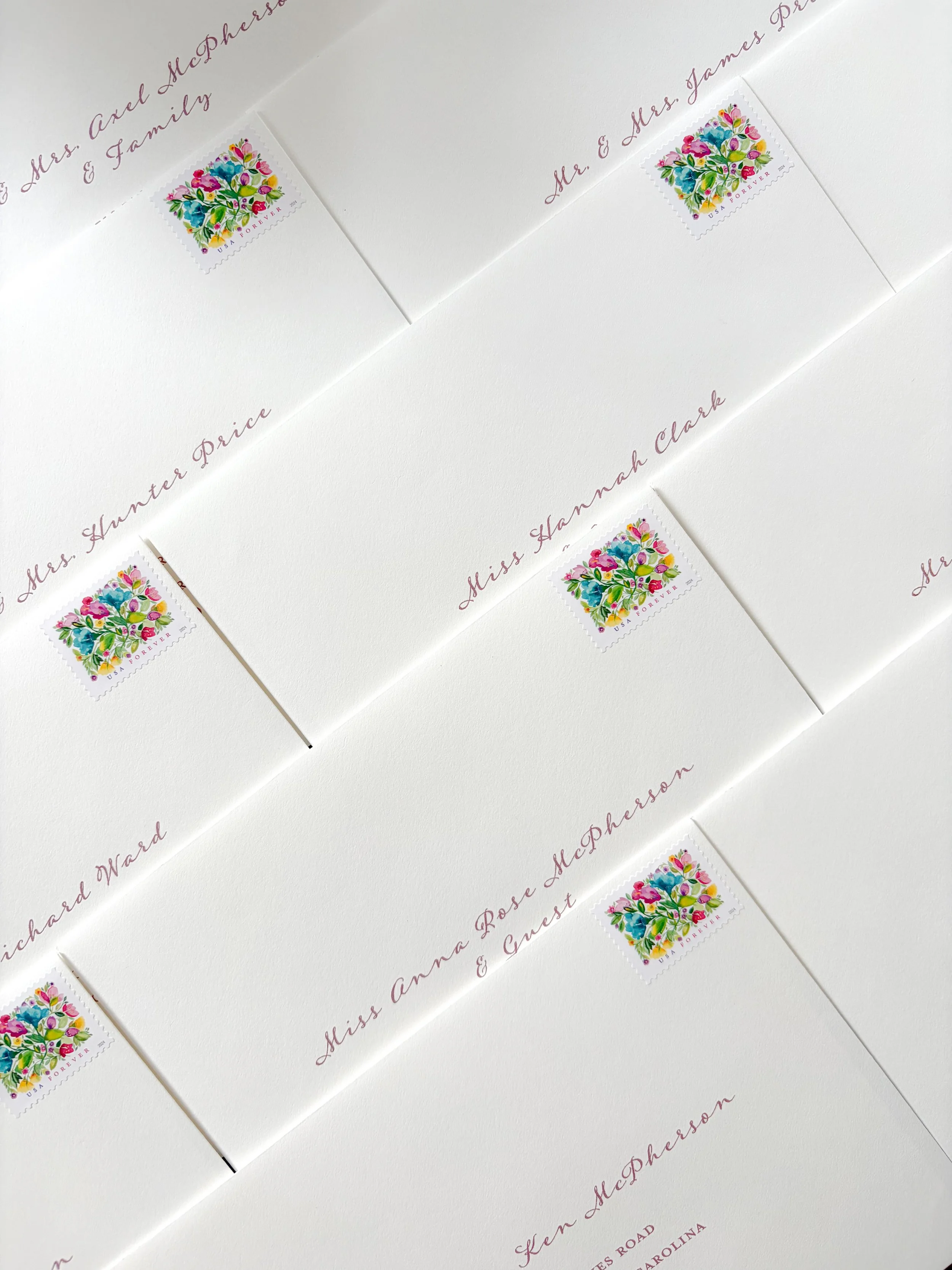 White envelopes with handwritten names in pink ink and colorful postage stamps featuring flowers, arranged diagonally on a white surface.