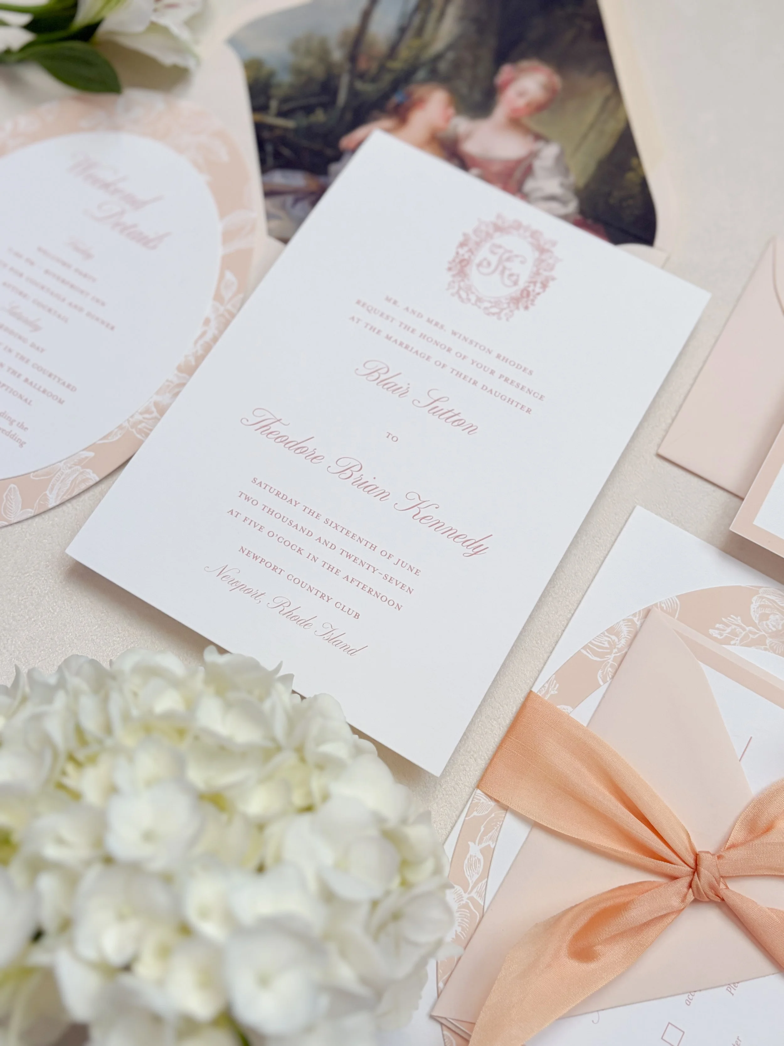 Elegant wedding invitation with blush pink accents, white floral decorations, and a peach-colored satin ribbon, placed on a light-colored surface with a bouquet of white flowers and an old painting in the background.