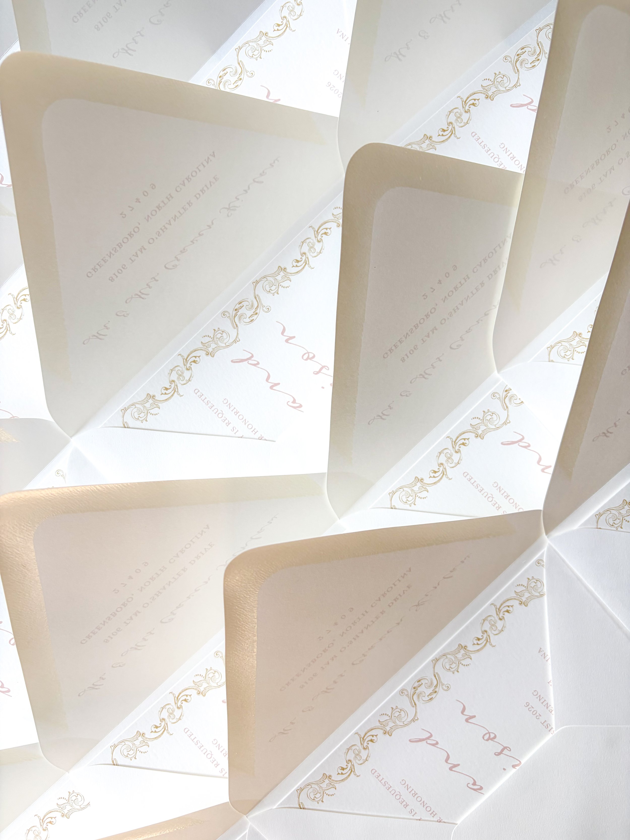 Multiple folded wedding invitations with gold ornate borders and light pink text on white paper, arranged in a fan shape.