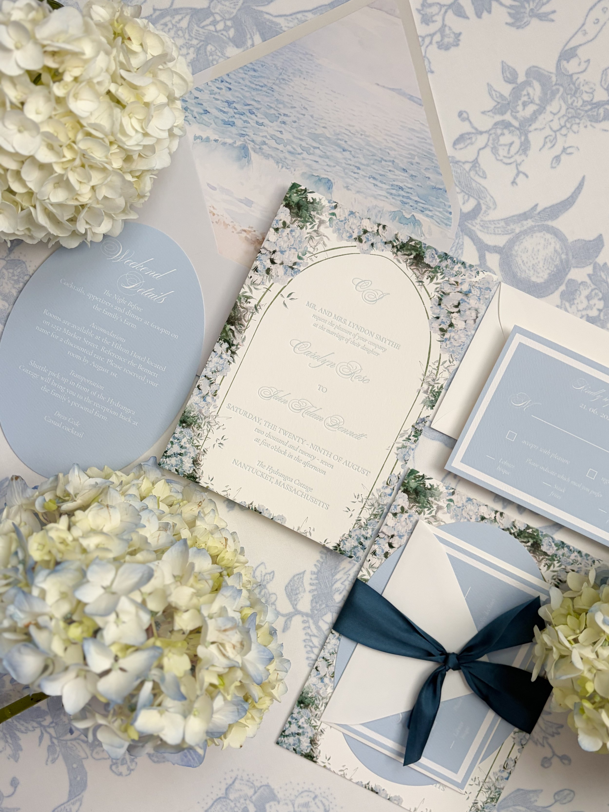 Wedding invitation suite with floral borders, surrounded by white hydrangeas, set on a blue patterned tablecloth.