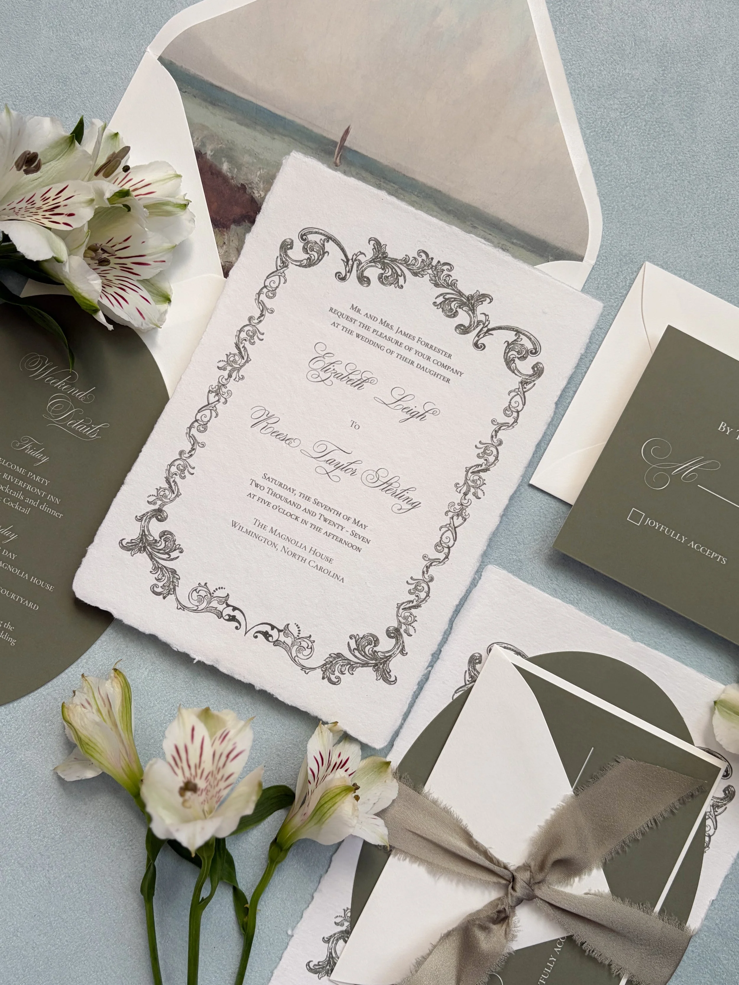 Invitation cards for a wedding with elegant floral and ornate design, accompanied by fresh white flowers with pink streaks and green leaves on a light blue surface.