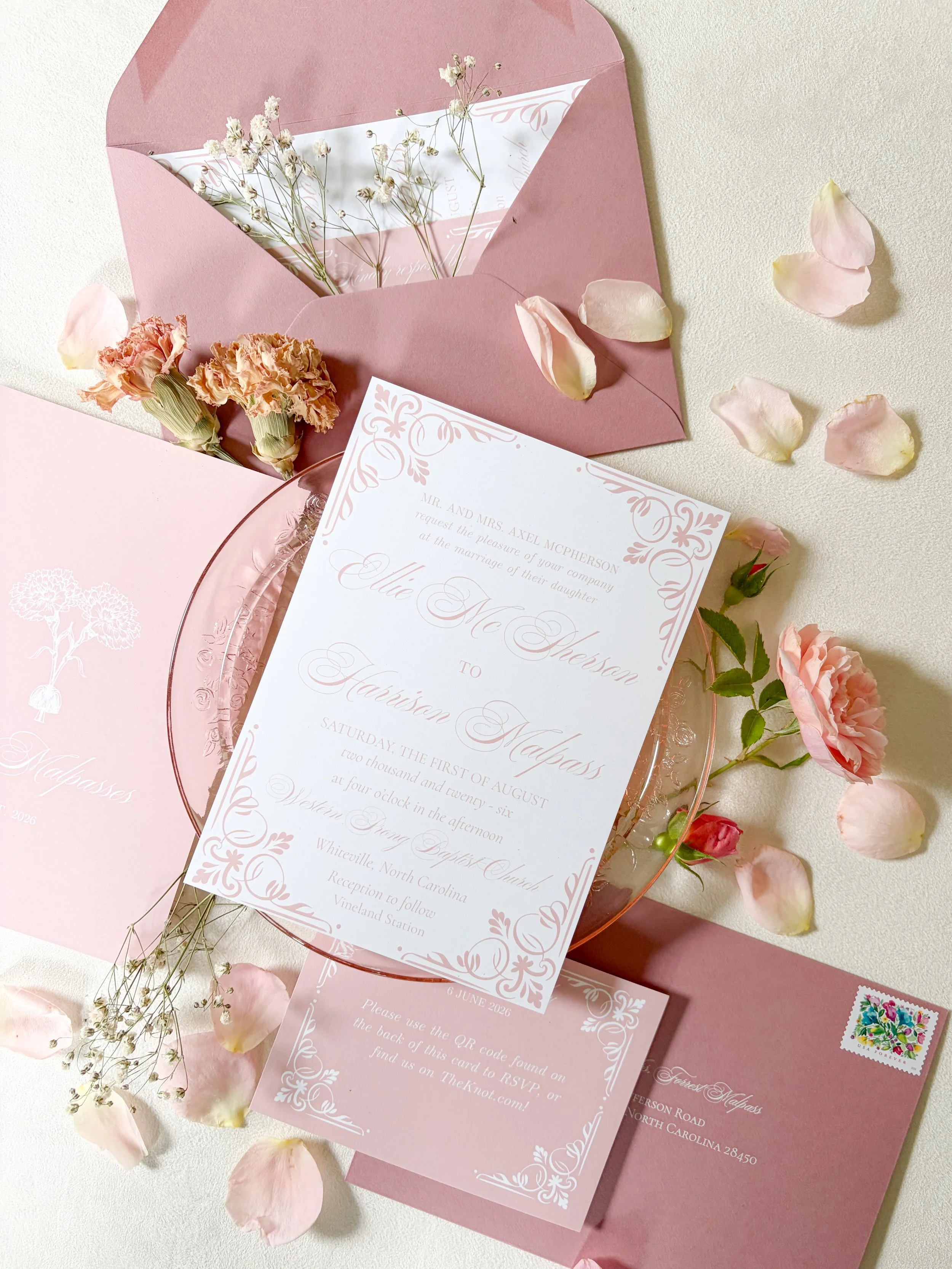 Pink wedding invitation suite with floral accents, including a printed card, pink envelope, and scattered pink rose petals and small flowers on a white background.