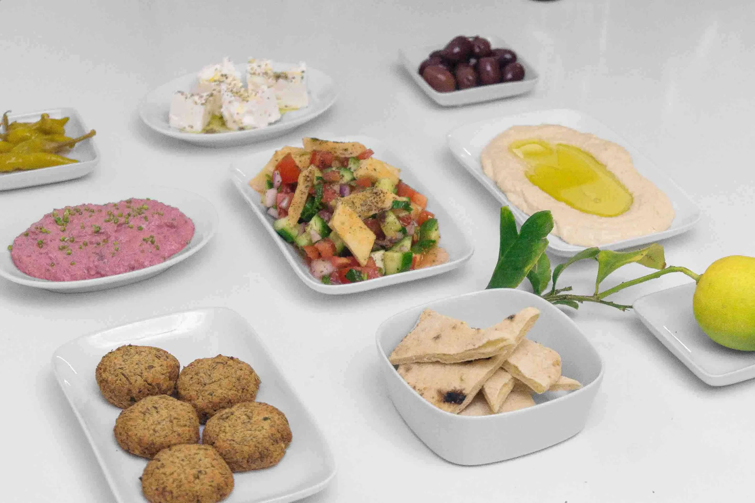 Various Middle Eastern dishes on white plates, including falafel, hummus with olive oil, tabbouleh salad, baba ganoush, stuffed grape leaves, and pickled vegetables, with a lemon and green leaves for garnish.