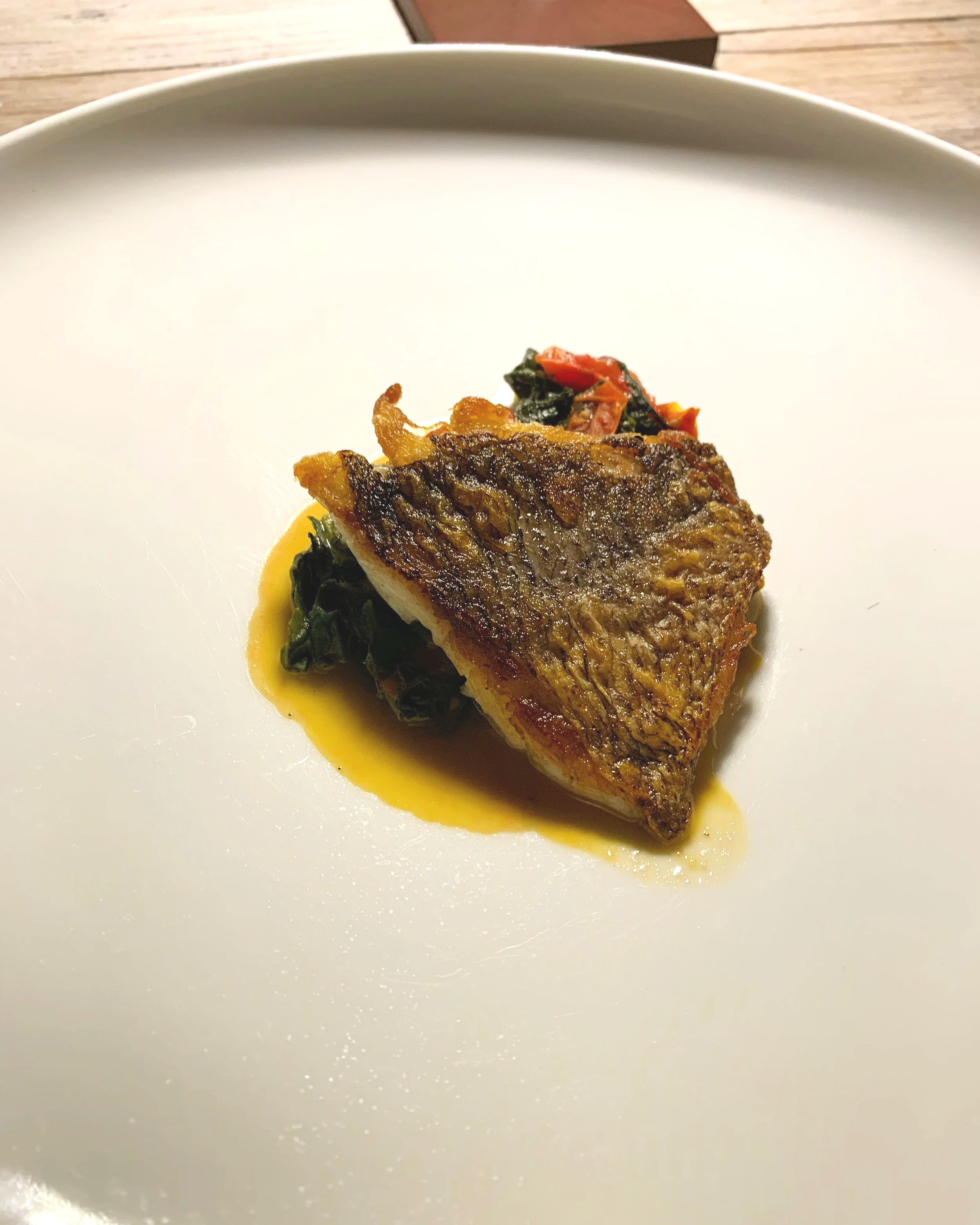A plated dish features a seared piece of fish with crispy skin, resting on a bed of cooked greens, and drizzled with a light, yellow sauce.