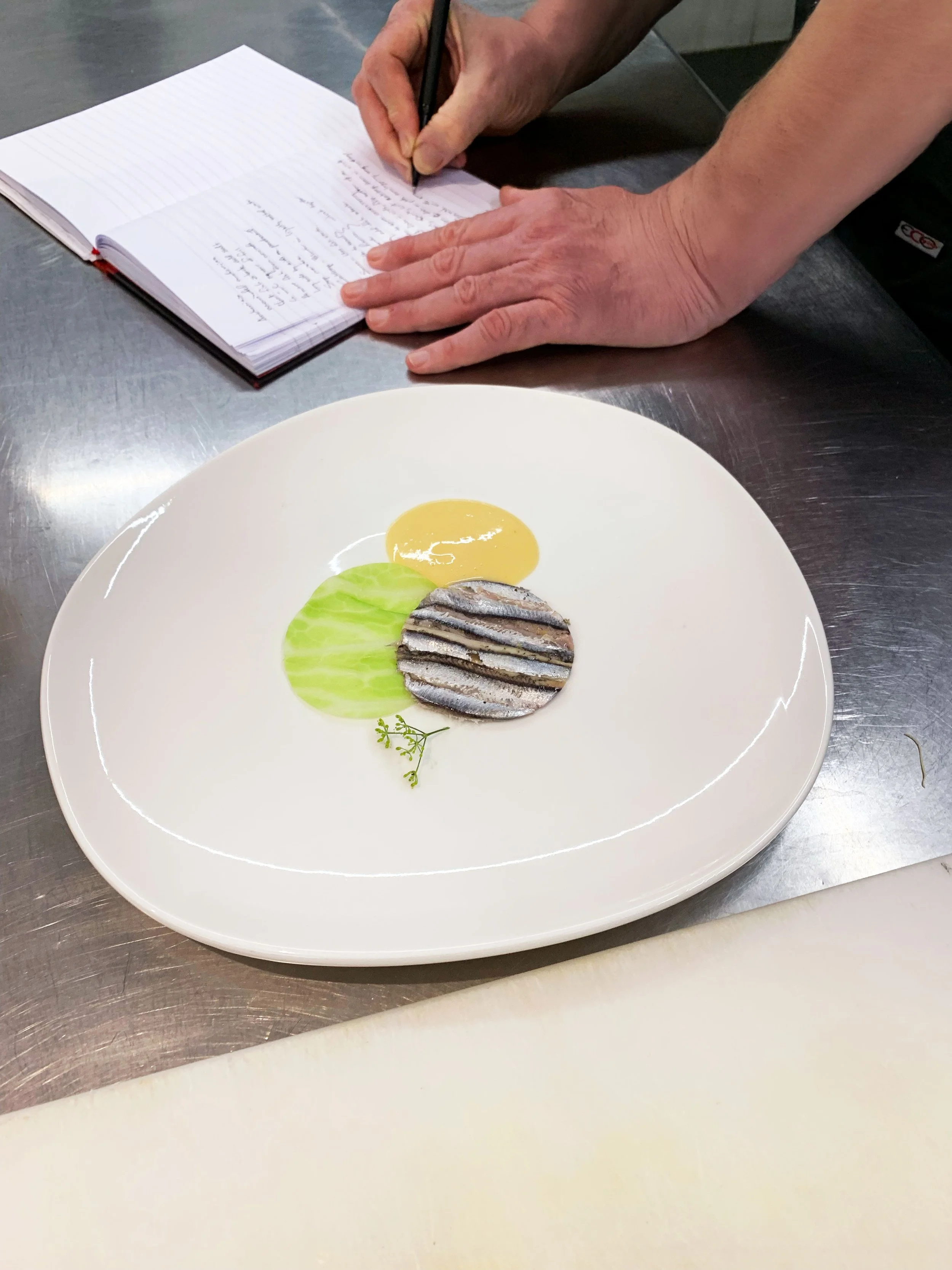 A chef preparing a dish with grilled fish served with thinly sliced vegetables, a yellow sauce, and a green sauce on a white plate, with a person writing on a notepad in the background.