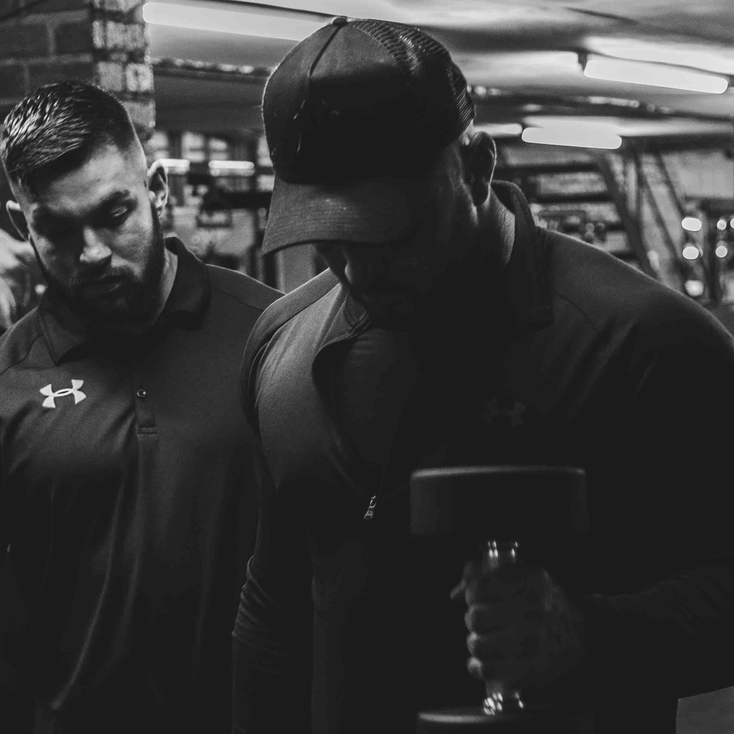 Two men in a gym, one holding a dumbbell, wearing Under Armour gear. Black and white image with low lighting.