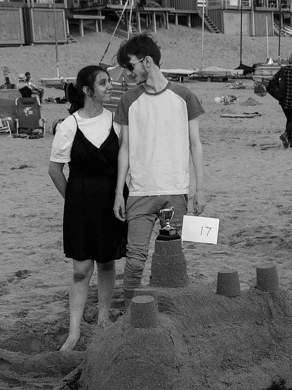 Two people on a beach standing next to a sandcastle with a trophy and a sign labeled "17" on it.