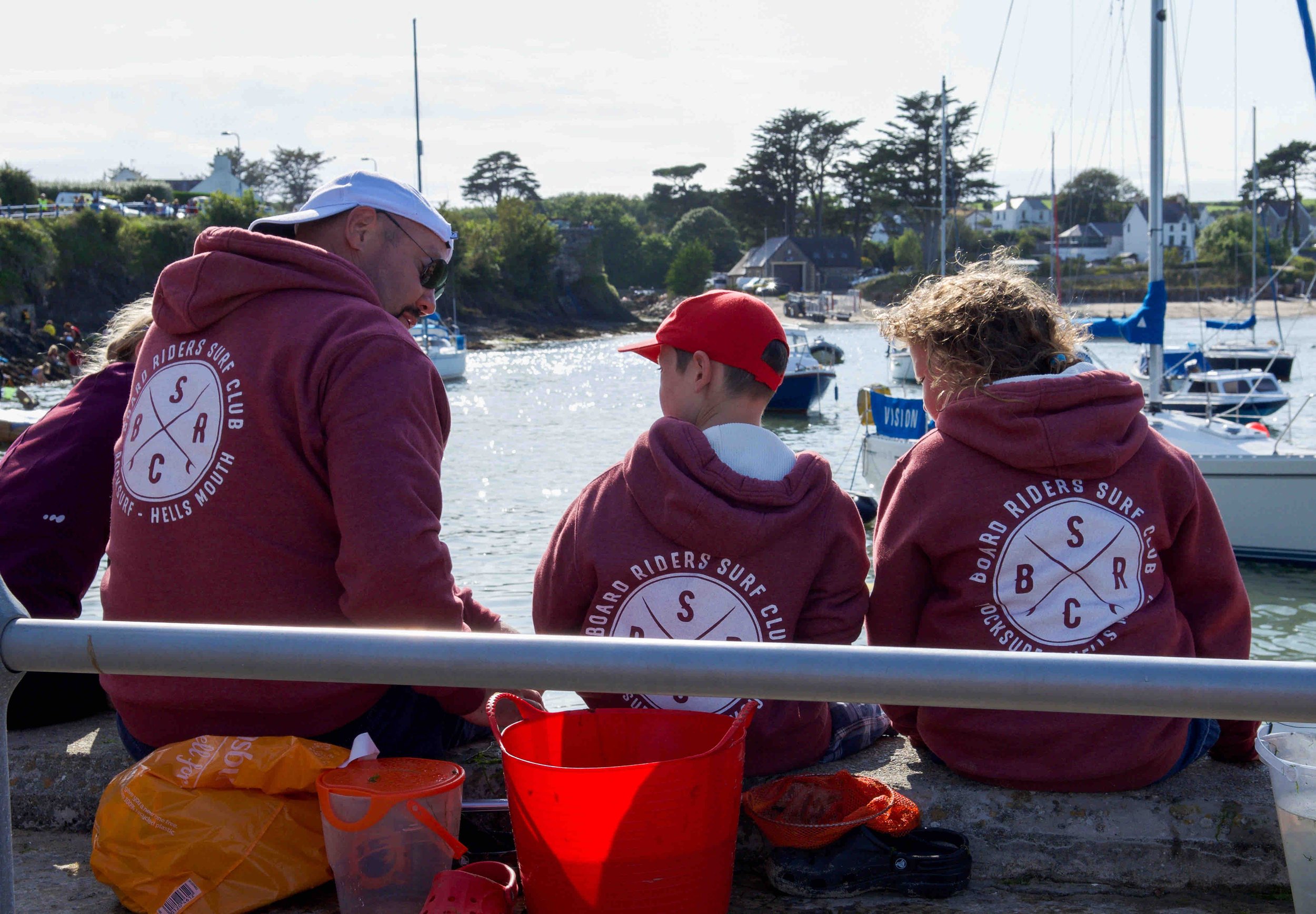 Group of three people wearing matching "Board Riders Surf Club" hoodies sitting by a harbor with boats in the background.