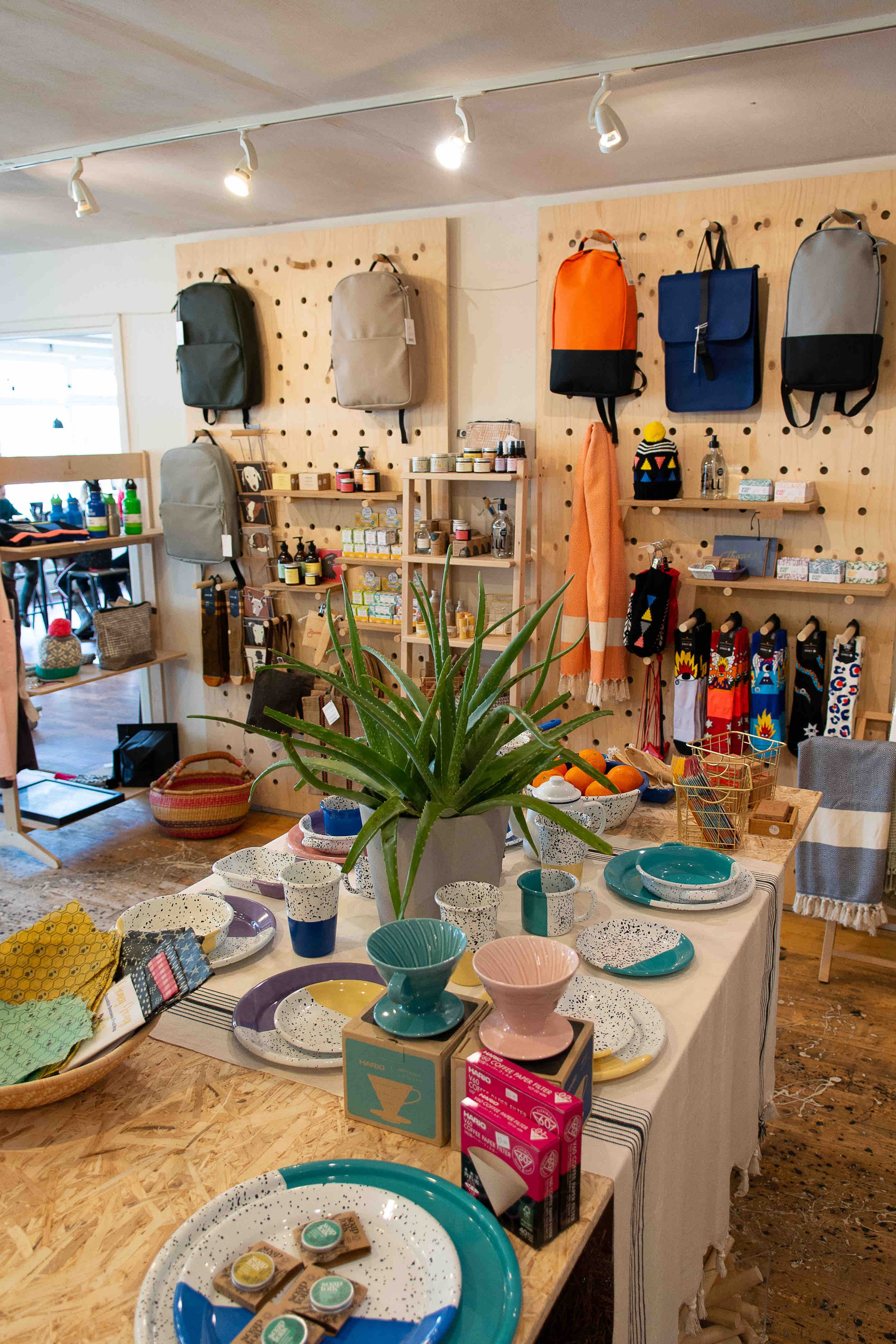 Retail store interior featuring a variety of products for sale. Items include backpacks, kitchenware, plant pots, and small home goods displayed on wooden shelves and tables. The scene is well-lit with track lighting, highlighting colorful ceramics,
