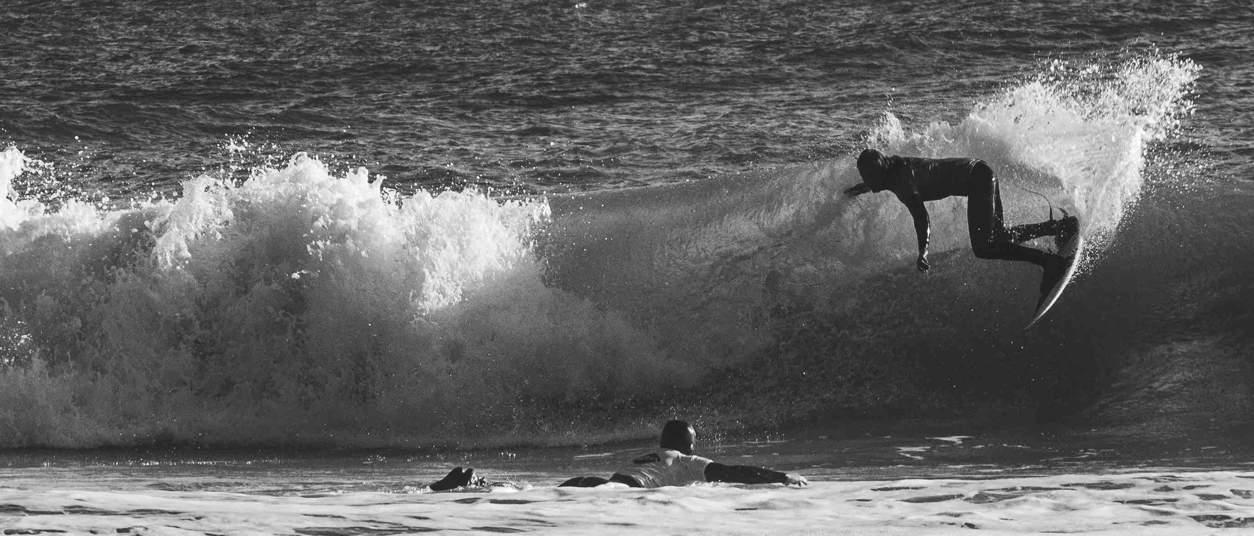 Black and white photo of a surfer riding a wave with another person watching from the water.