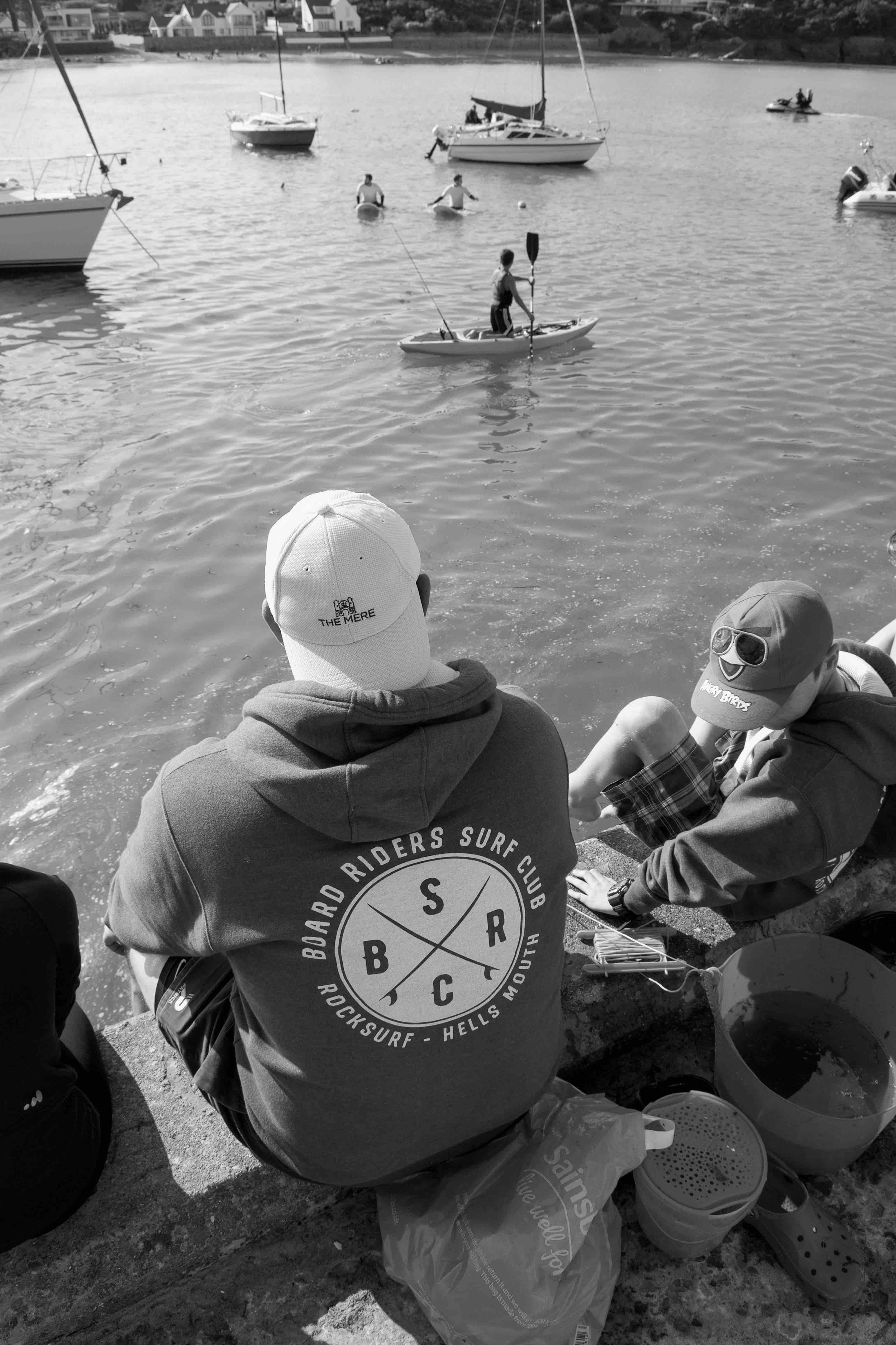 People sitting on a pier by water with boats and kayakers nearby.