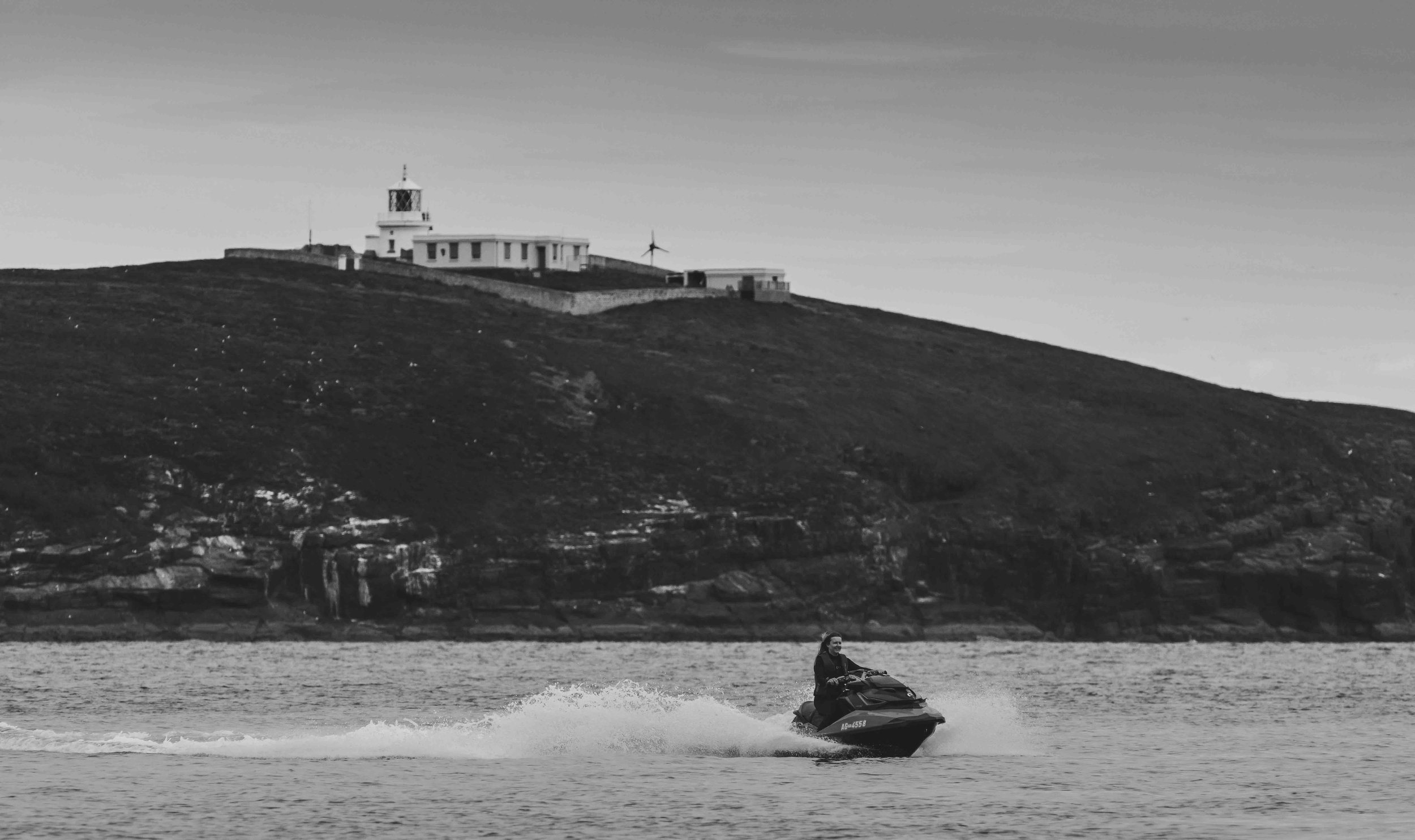 Person riding a jet ski on water in front of a rocky hill with a lighthouse on top.