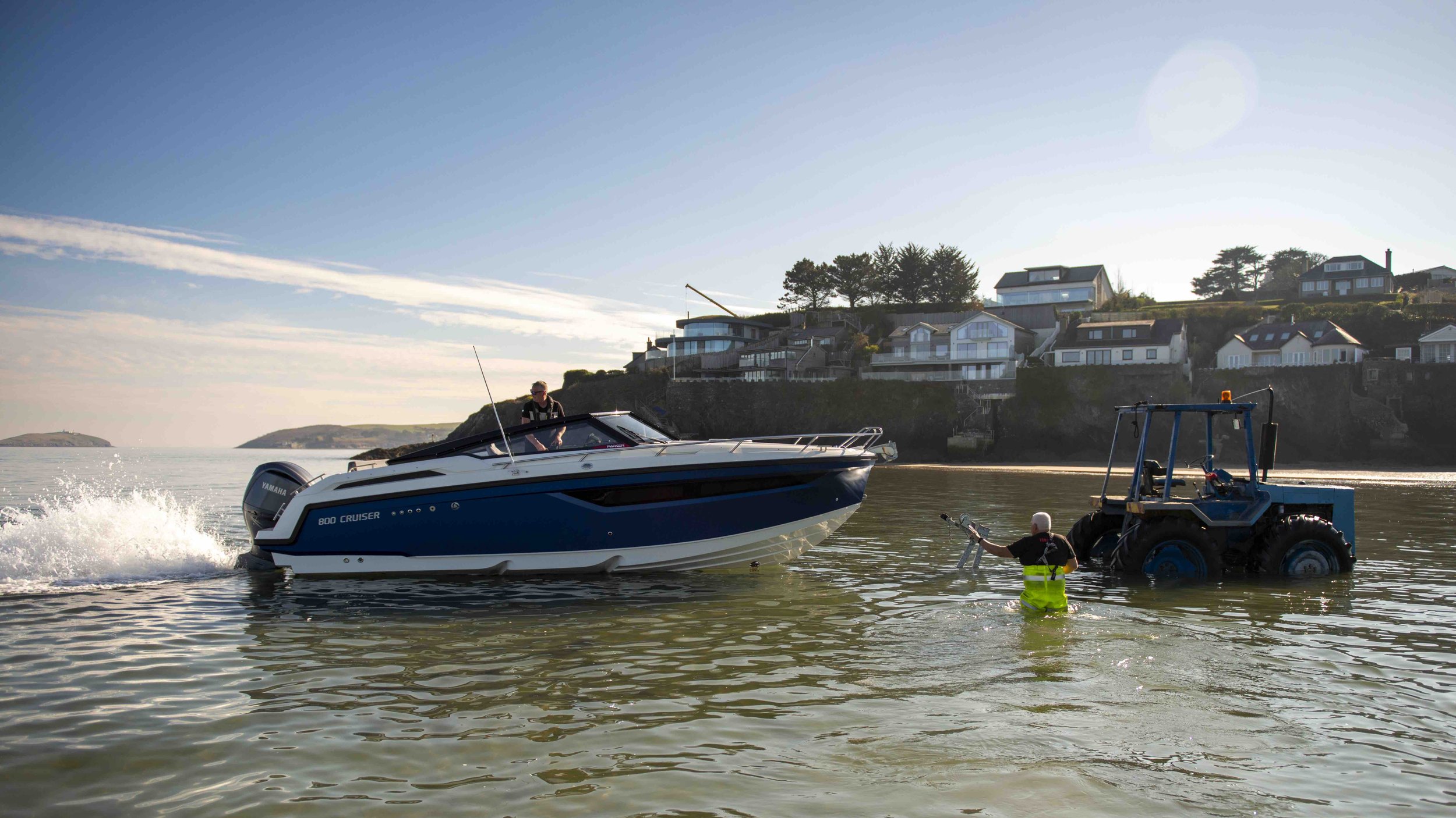 Motorboat being launched into the water by a tractor on a beach with houses on a cliff in the background.