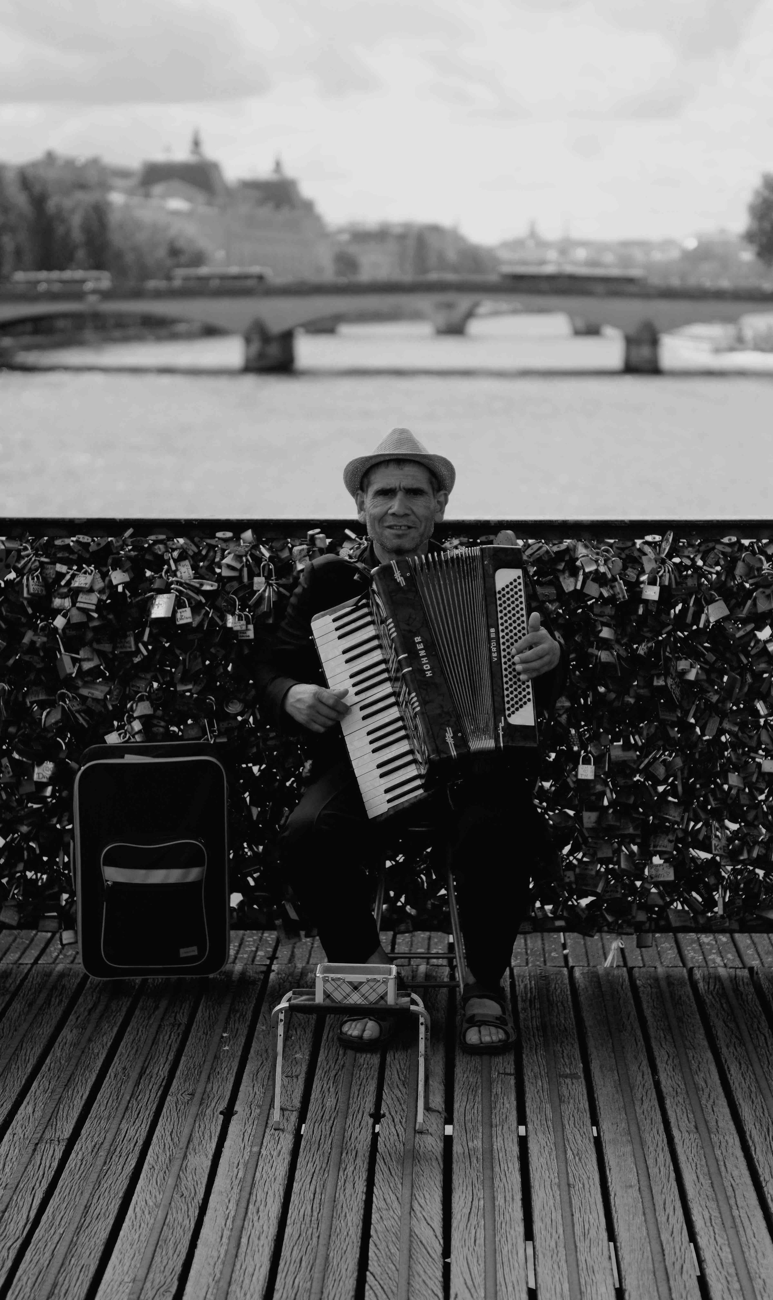 Man playing an accordion on a wooden bridge with a background of a river and a bridge with padlocks.