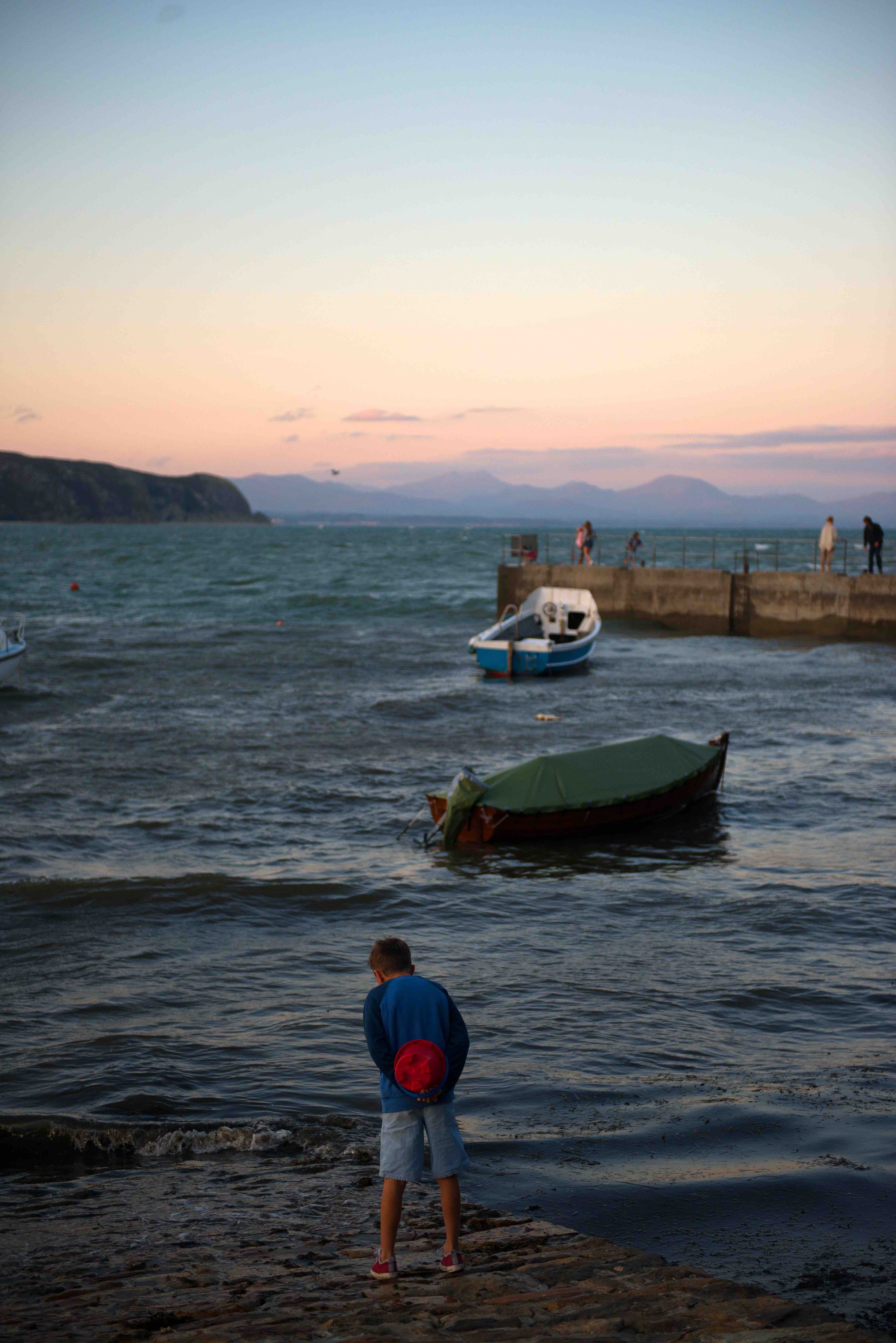 Boy on a rocky shoreline looking at a boat on the water, with a pier and hills in the background during sunset.