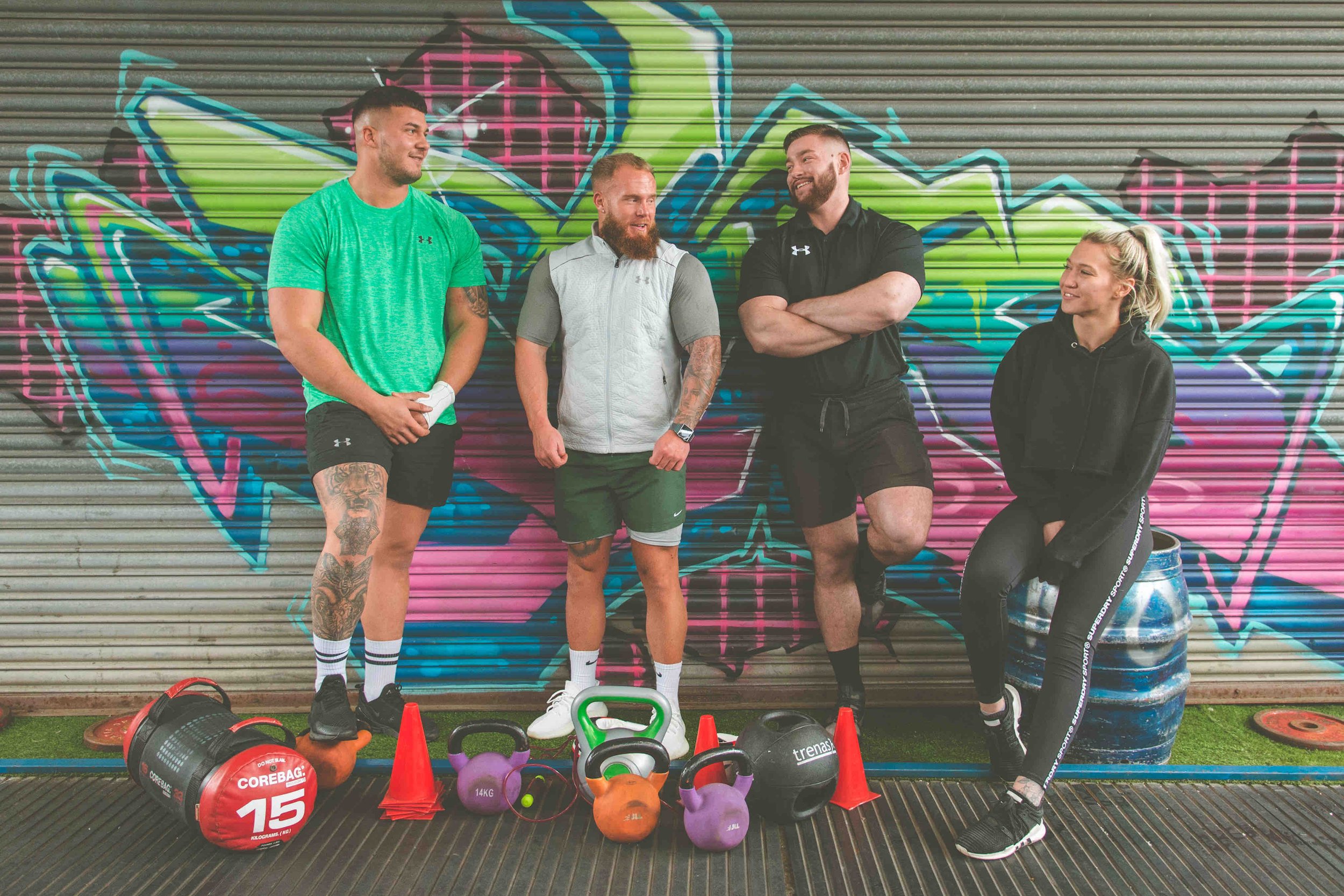 Group of four people in gym setting with kettlebells and graffiti background