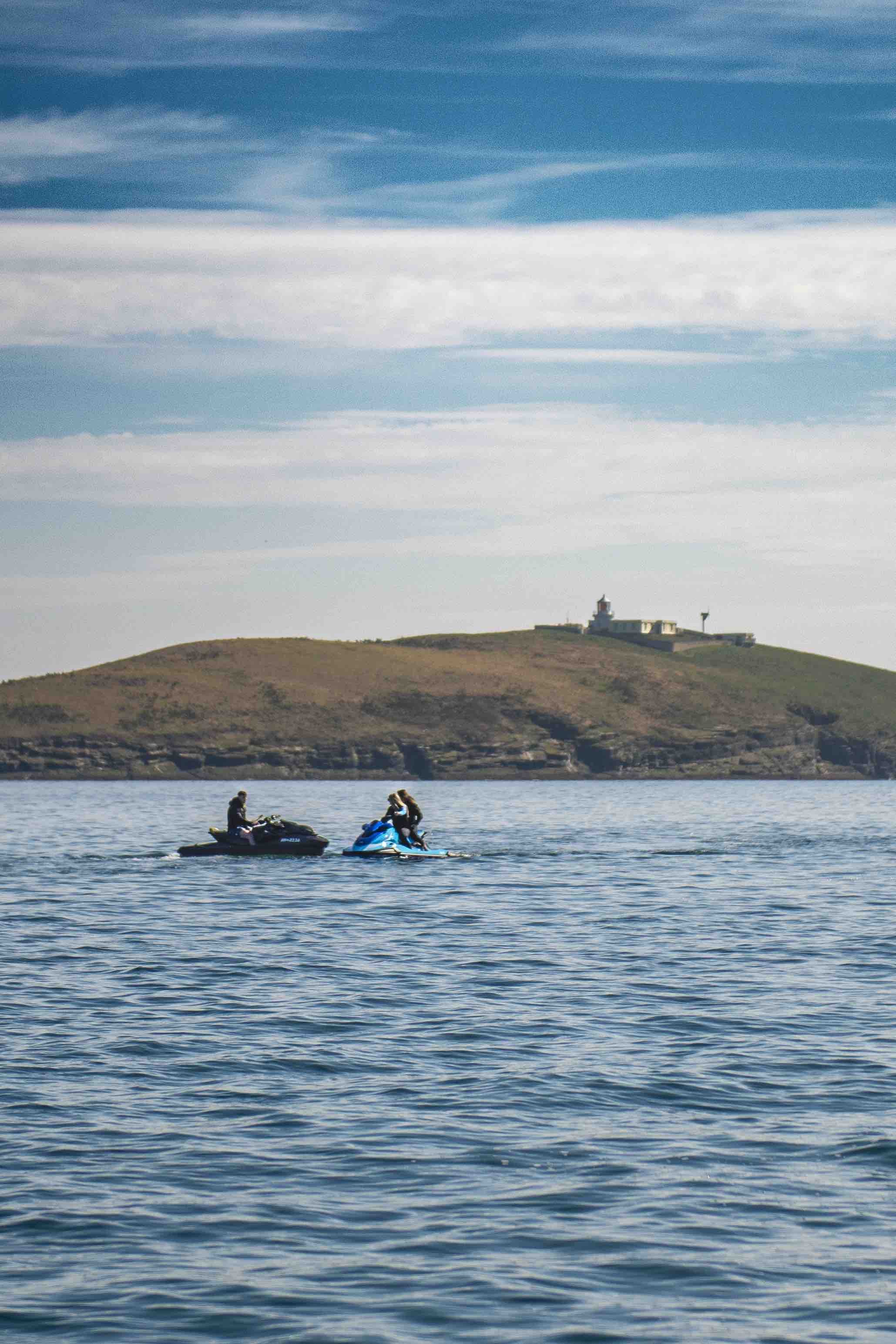 Two people riding jet skis on the ocean near a small island with a building on it under a partly cloudy sky.