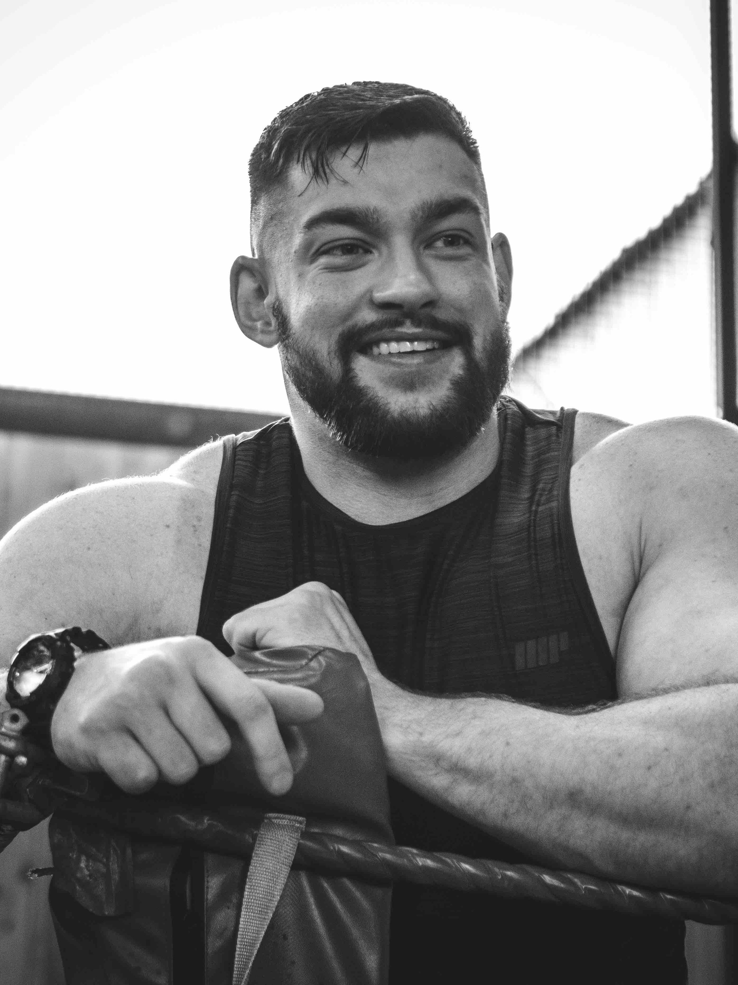 Black and white photo of a smiling person with a beard, leaning on a padded surface, wearing a sleeveless shirt and a wristwatch.