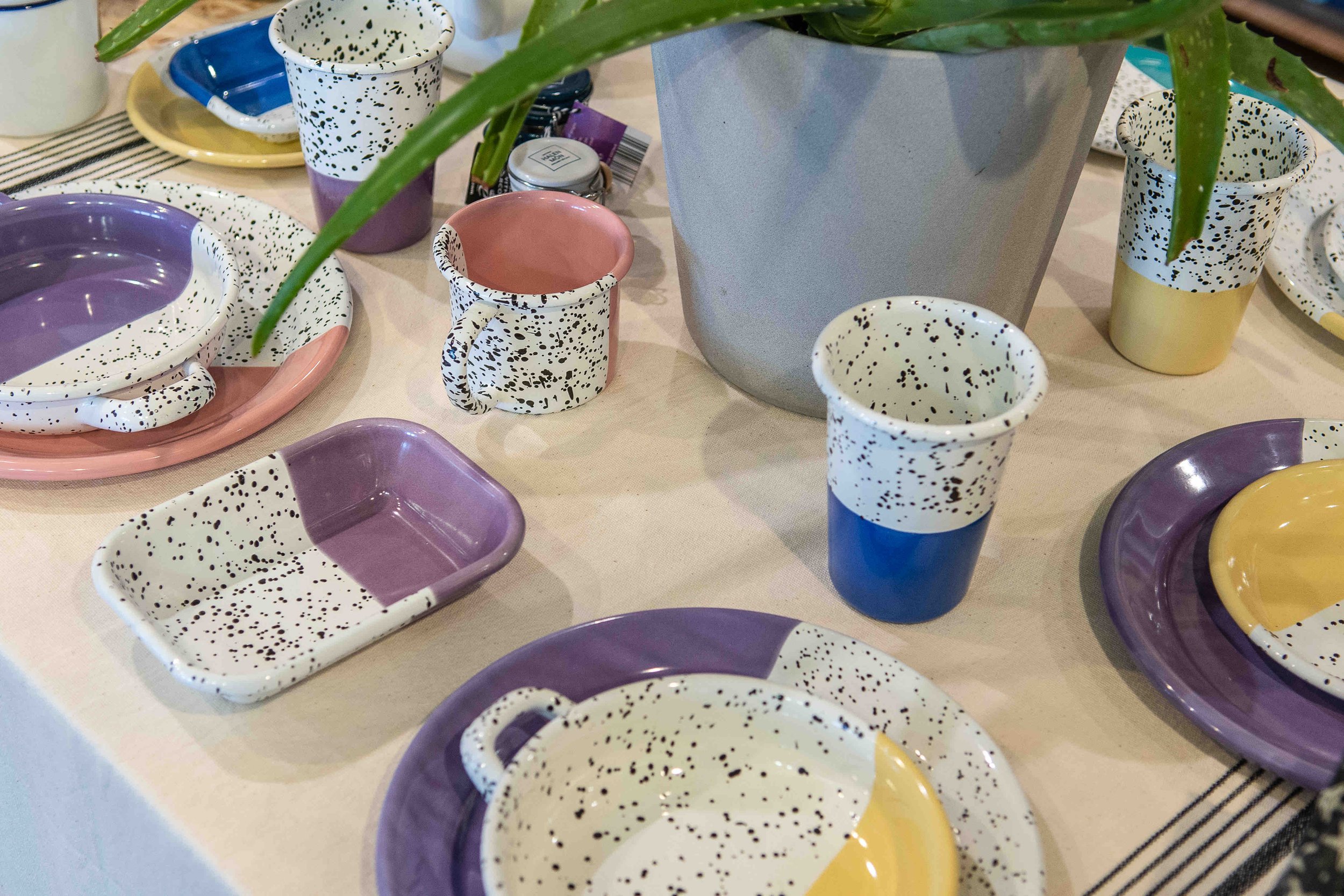 A table setting with colorful, speckled ceramic dishes, including plates, cups, and pitchers. The table also features a gray plant pot with green leaves. The ceramics have a mix of pastel hues like purple, pink, yellow, and blue, combined with black