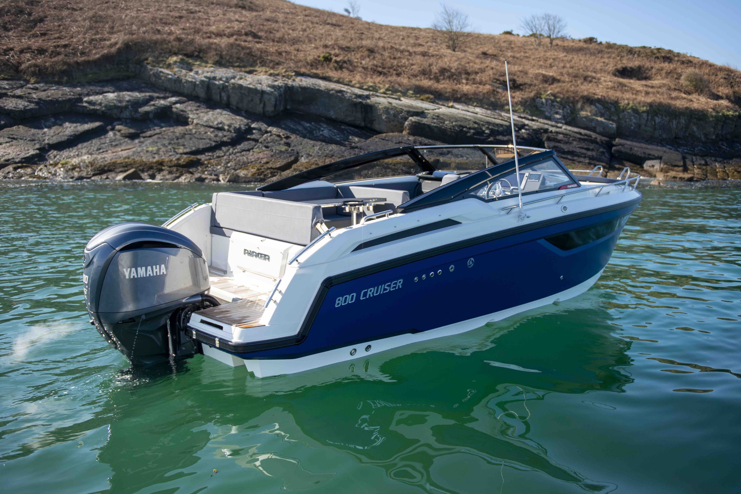A blue and white Parker 800 Cruiser boat with a Yamaha outboard motor is on calm water near a rocky shoreline.