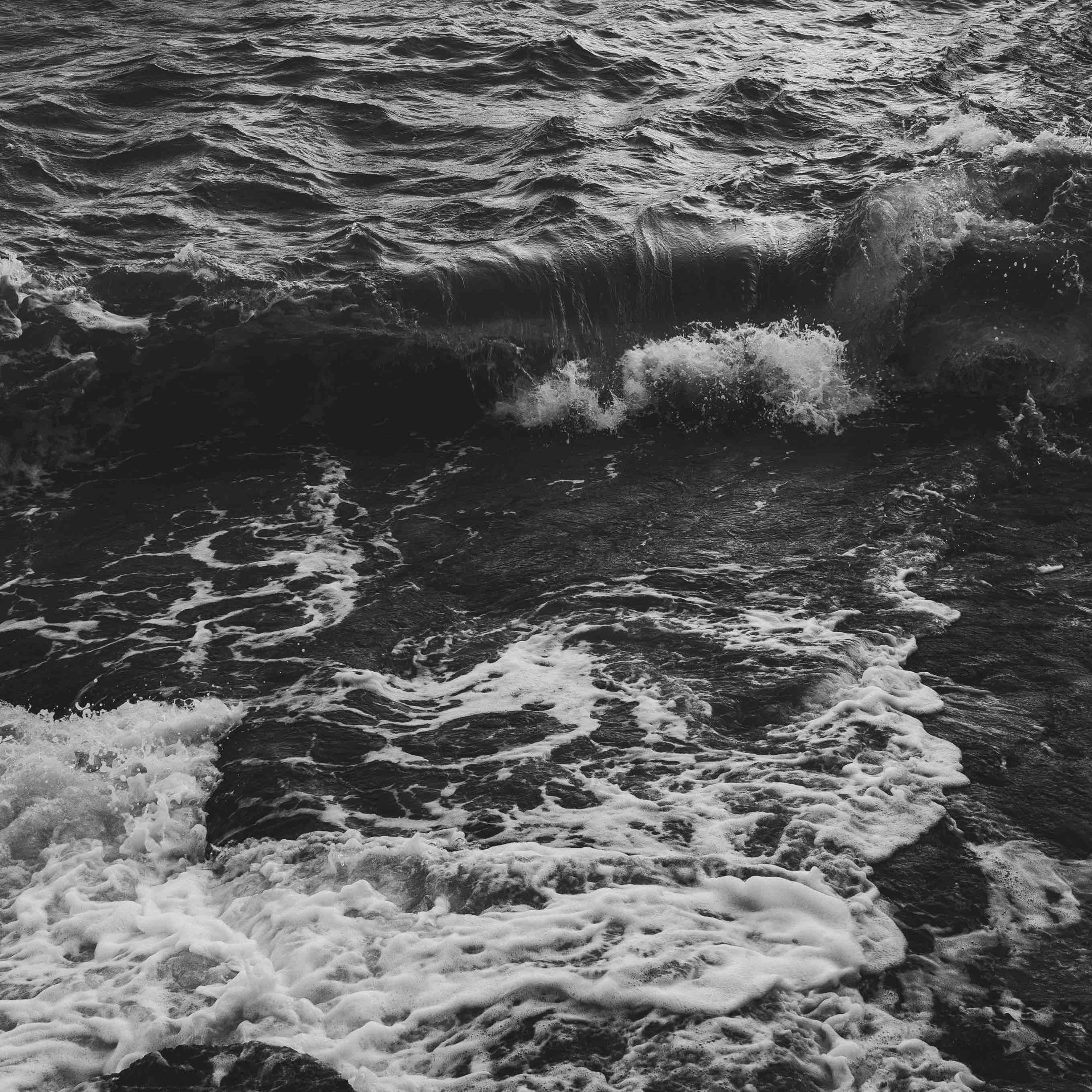 Black and white photo of ocean waves crashing on rocks, with white foam and ripples.