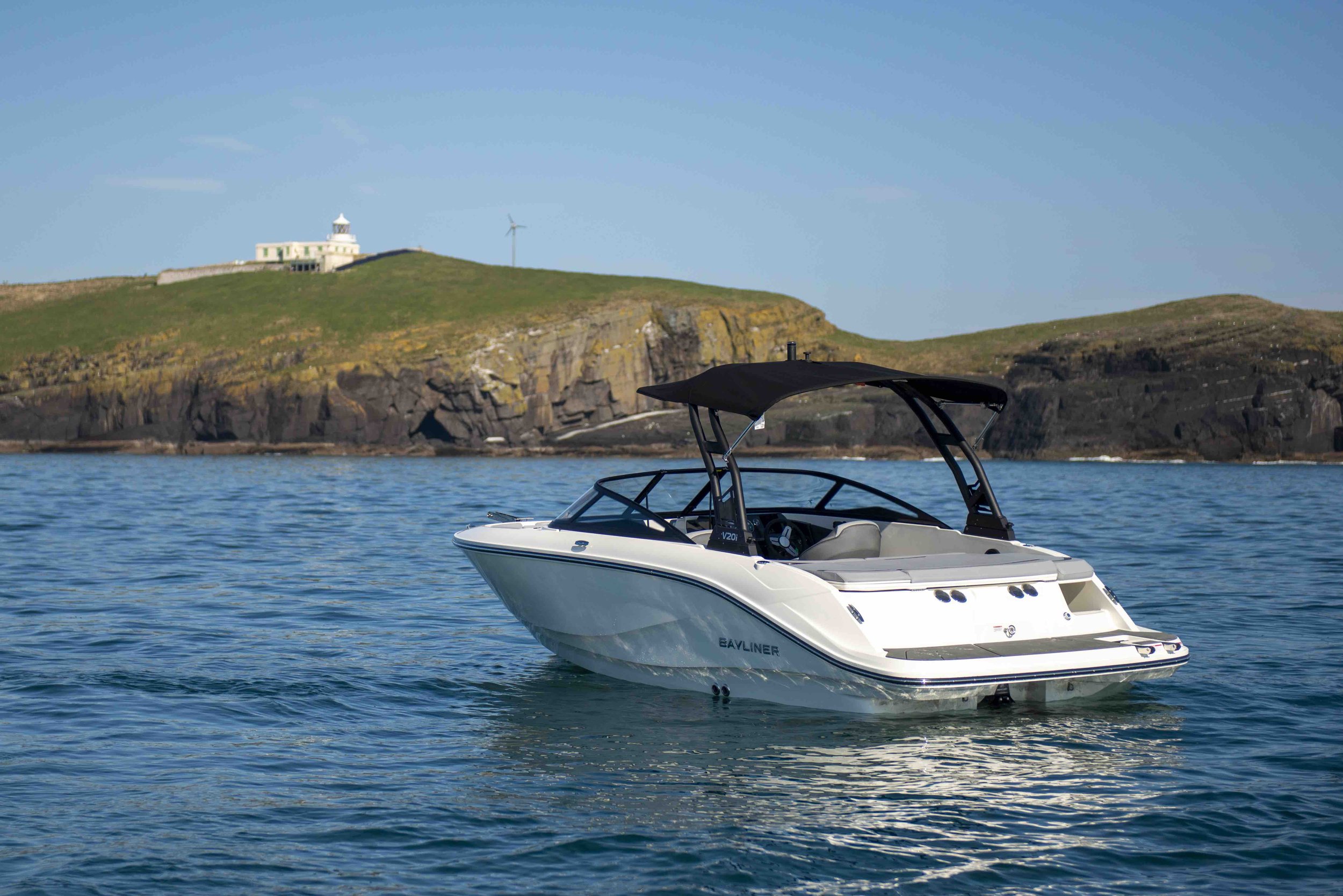 Speedboat on the water near rocky coastline with green hills and a lighthouse in the background.