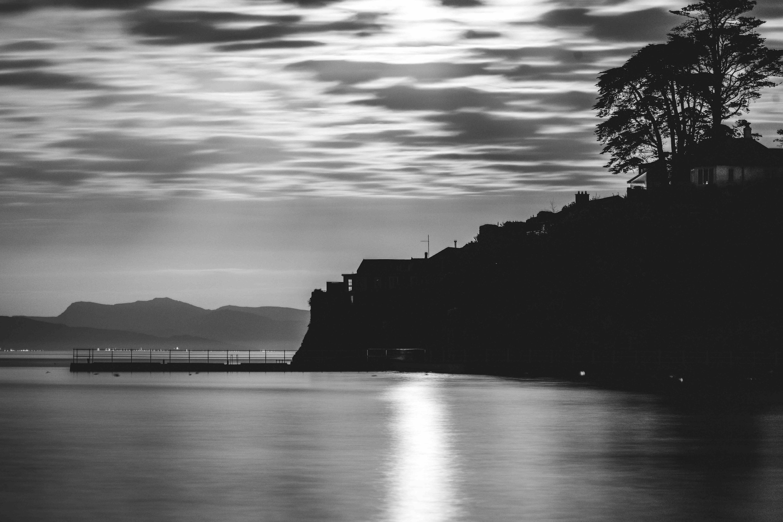 Black and white photo of a coastal landscape with silhouetted buildings and trees against a cloudy sky, overlooking calm water reflecting light.