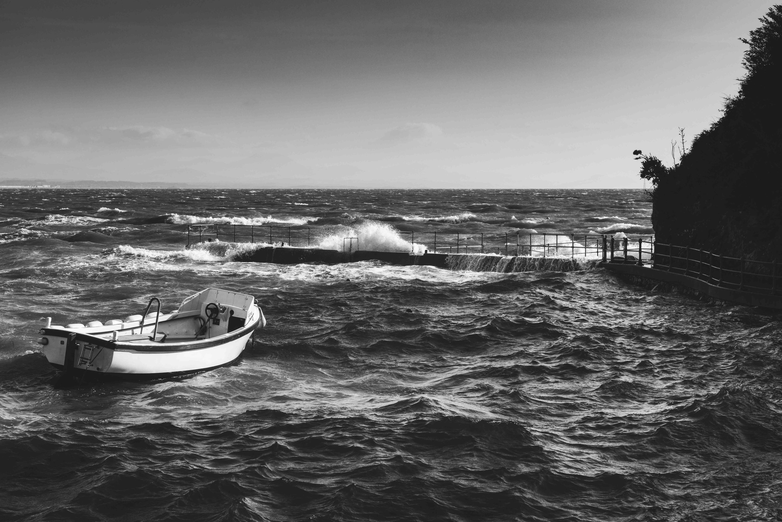 Black and white photo of a small boat in rough sea waters near a rocky shoreline with crashing waves.