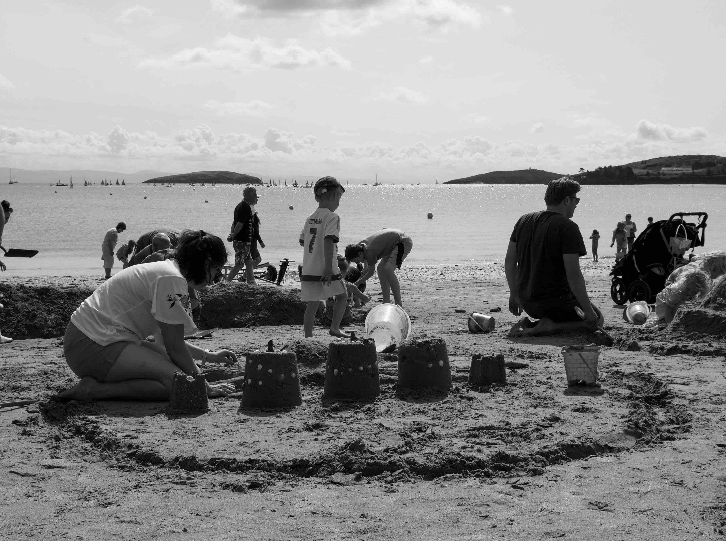 People building sandcastles on a beach with the sea and sailboats in the background.