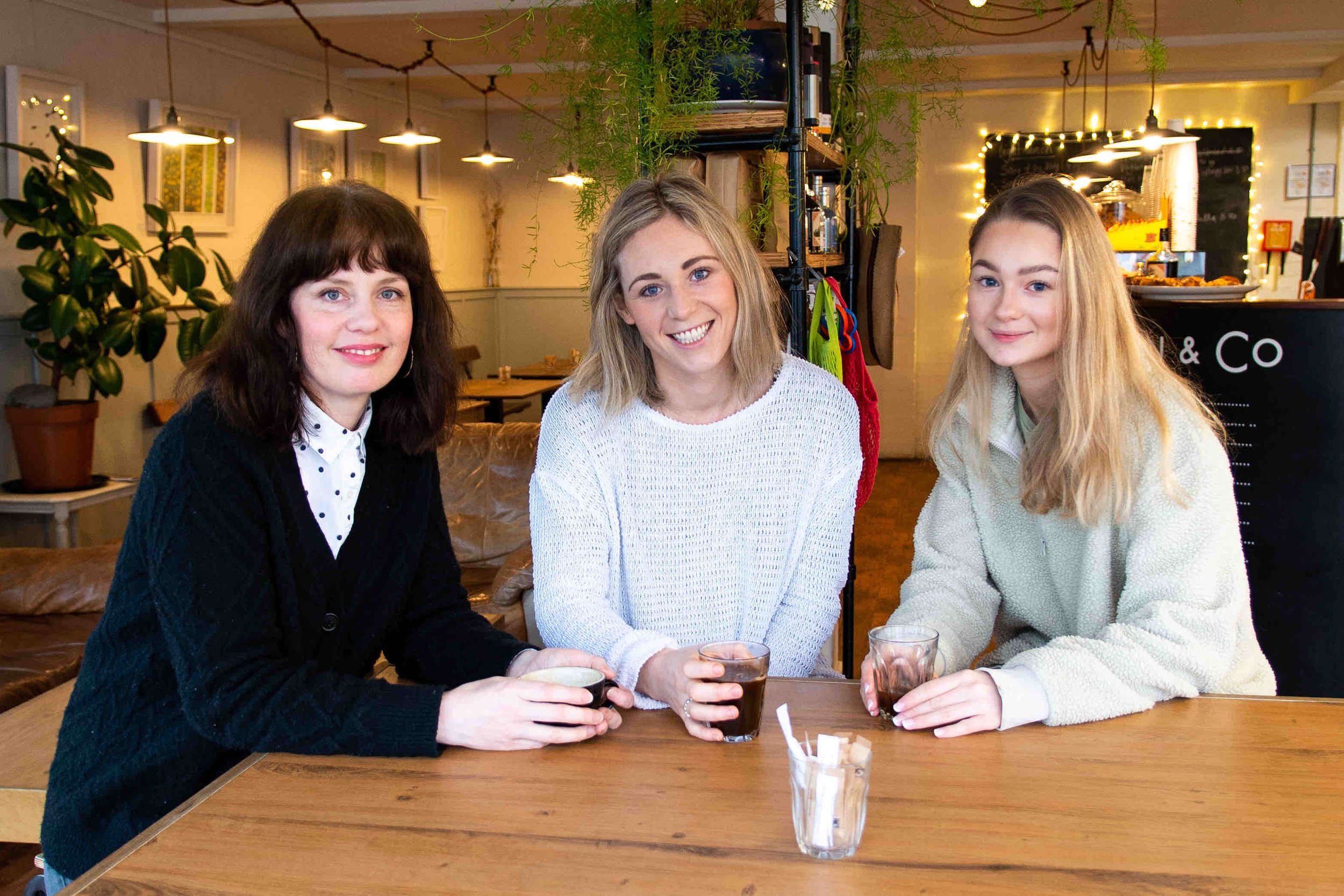 Three women sitting at a wooden table in a cozy café, holding drinks and smiling, with plants and hanging lights in the background.