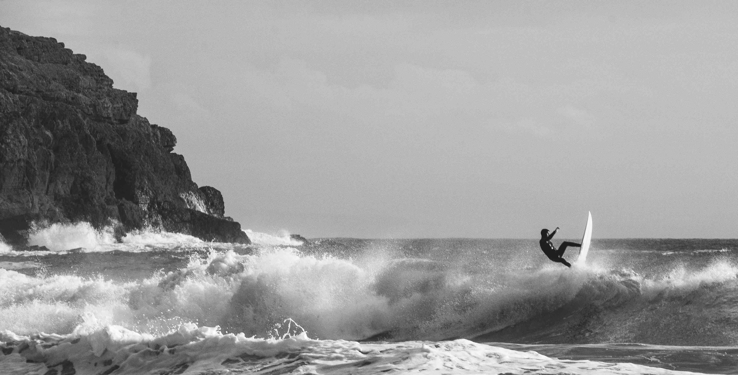 Black and white photo of a surfer performing a trick on a wave near cliffs.