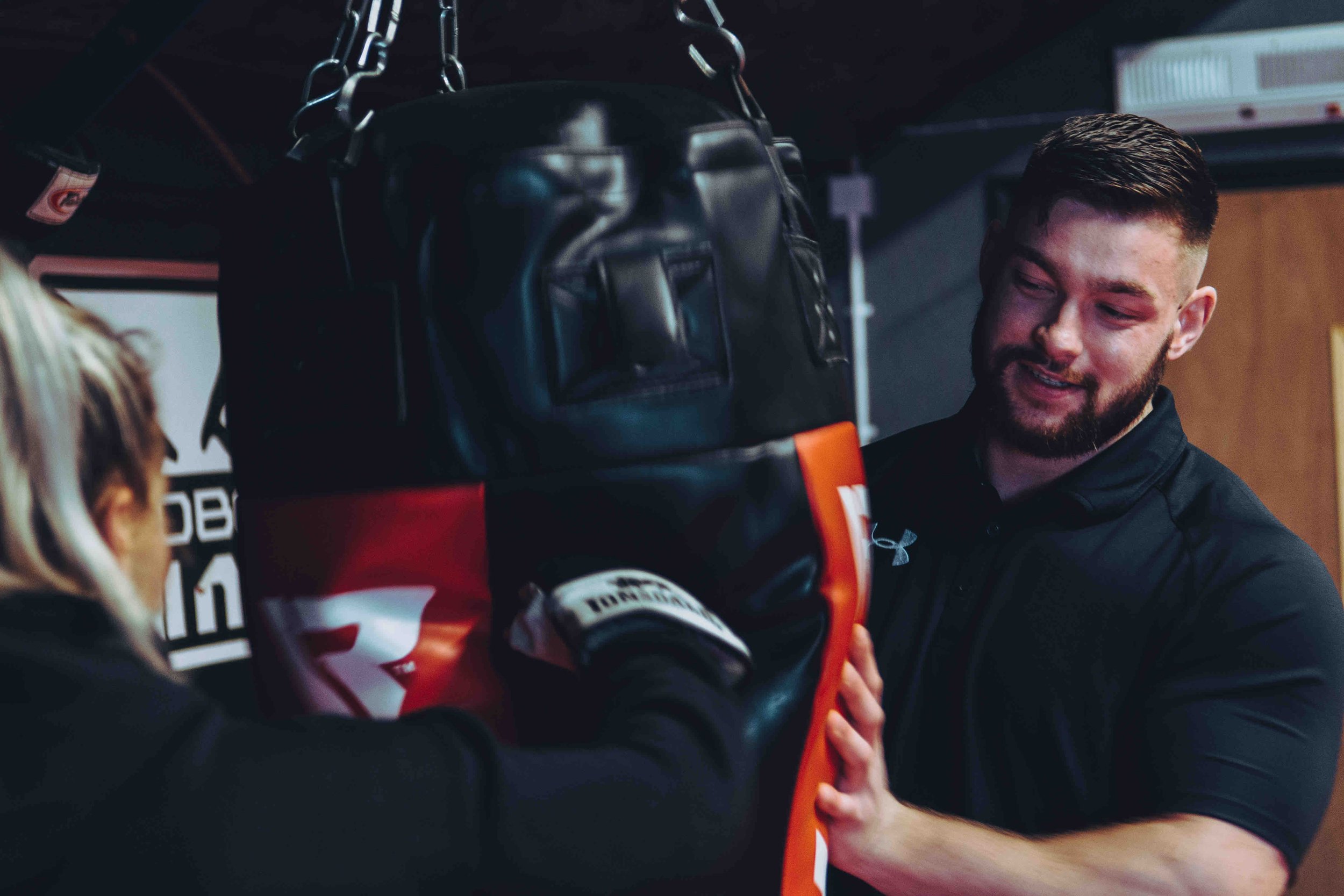 Two people training with a punching bag, indoors, wearing athletic clothing.