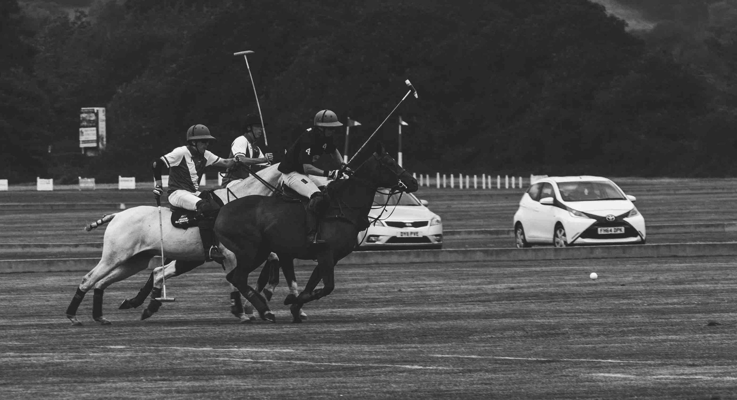 Black and white photo of a polo match in action, featuring two players on horseback, holding mallets, on a grass field. A white car is visible in the background.