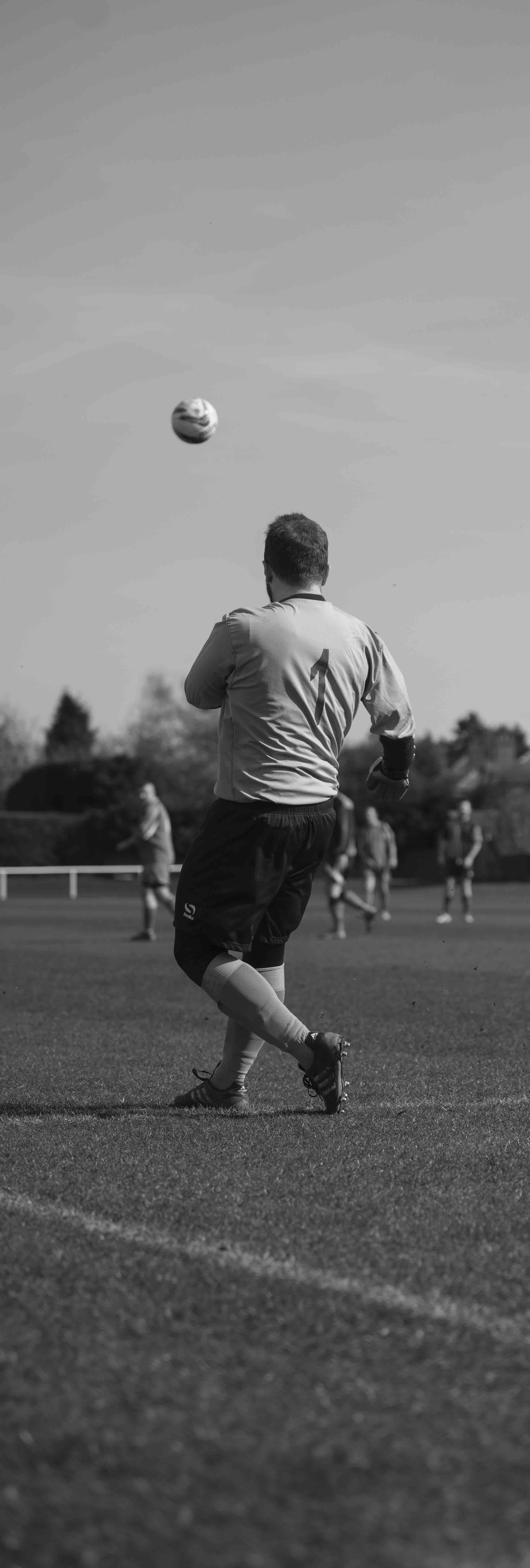 Black and white image of a soccer goalkeeper wearing the number 1 jersey, about to kick a soccer ball on a field. Several players are visible in the background.