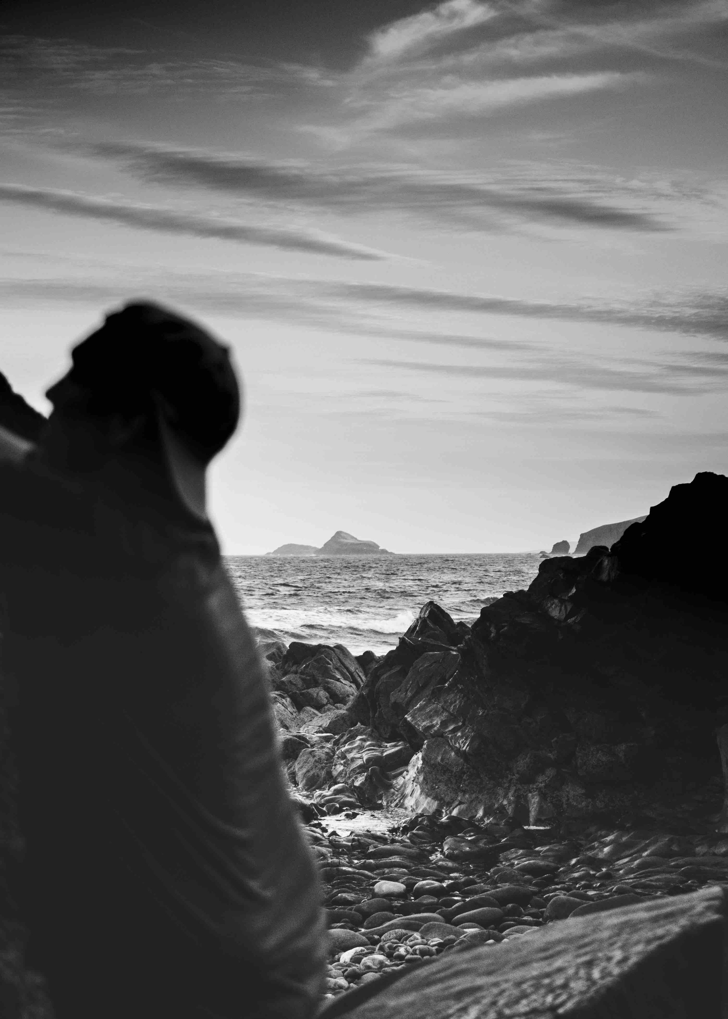 Black and white photo of a rocky beach with an ocean view, featuring a silhouette of a person in the foreground and distant islands on the horizon.