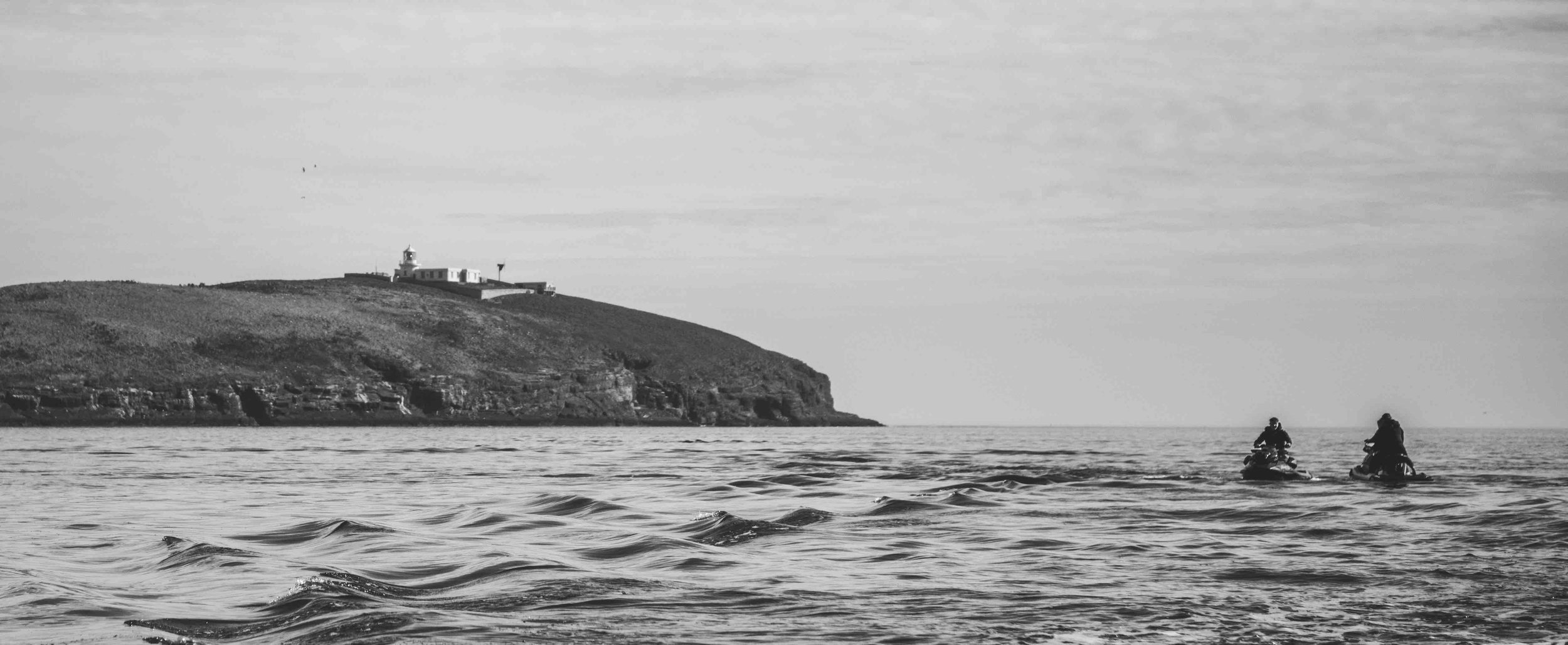 Two people on jet skis approaching an island with a lighthouse, black and white photo