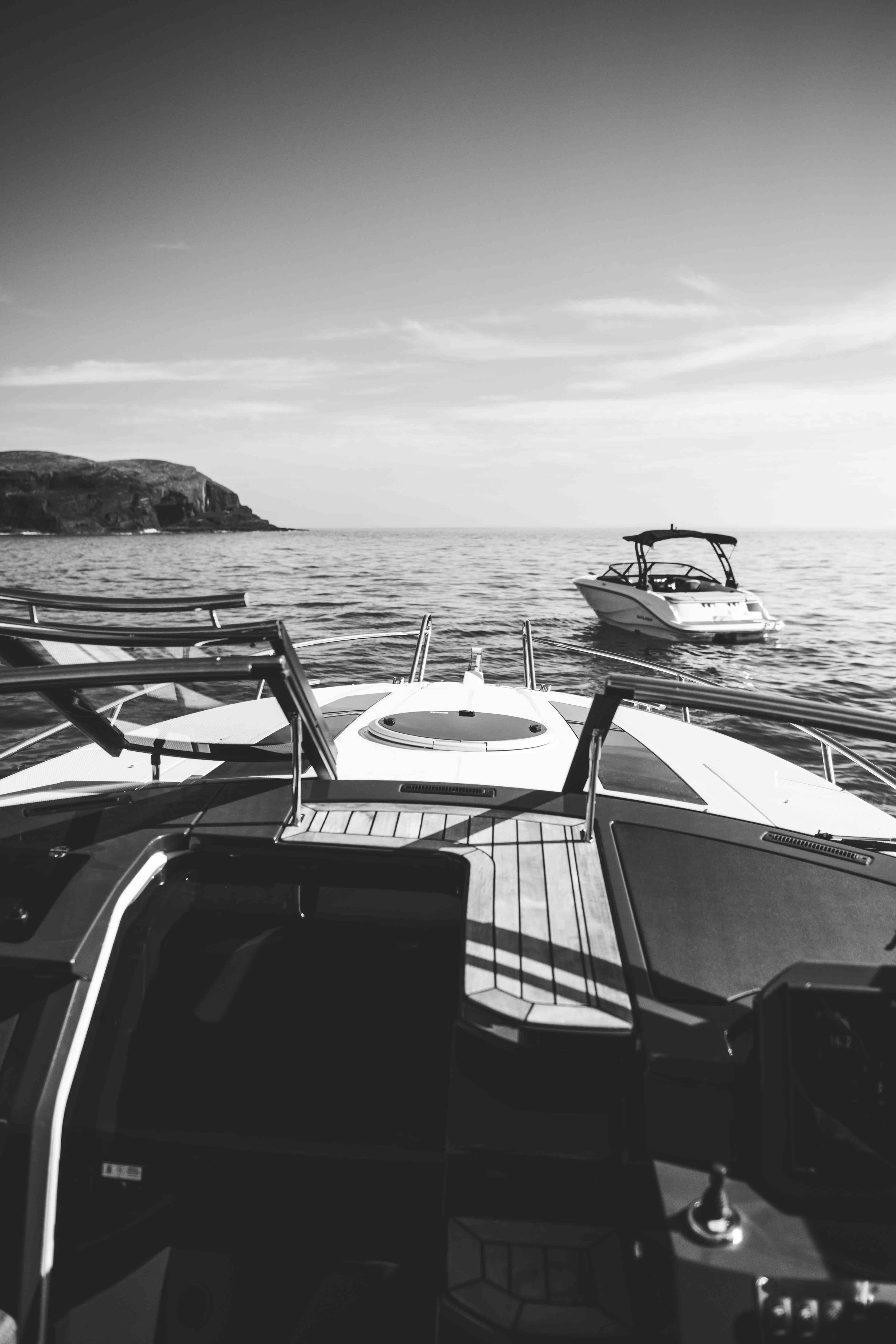 Black and white photo of two boats on the ocean, view from one boat's deck towards another boat, with a cliff in the background.