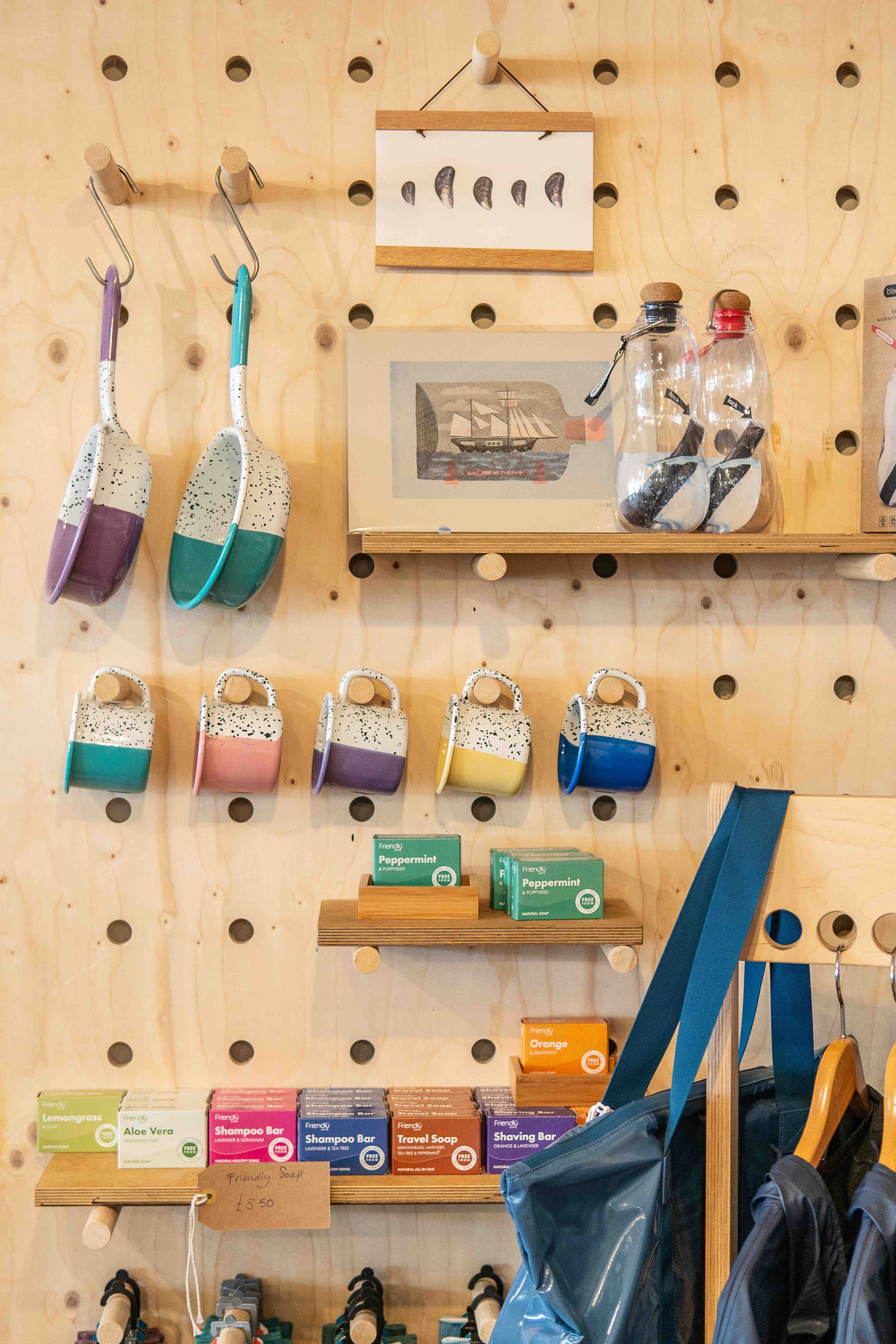 Wooden pegboard display with hanging kitchen utensils and mugs, framed art, reusable bottles, and shelves with soap products.