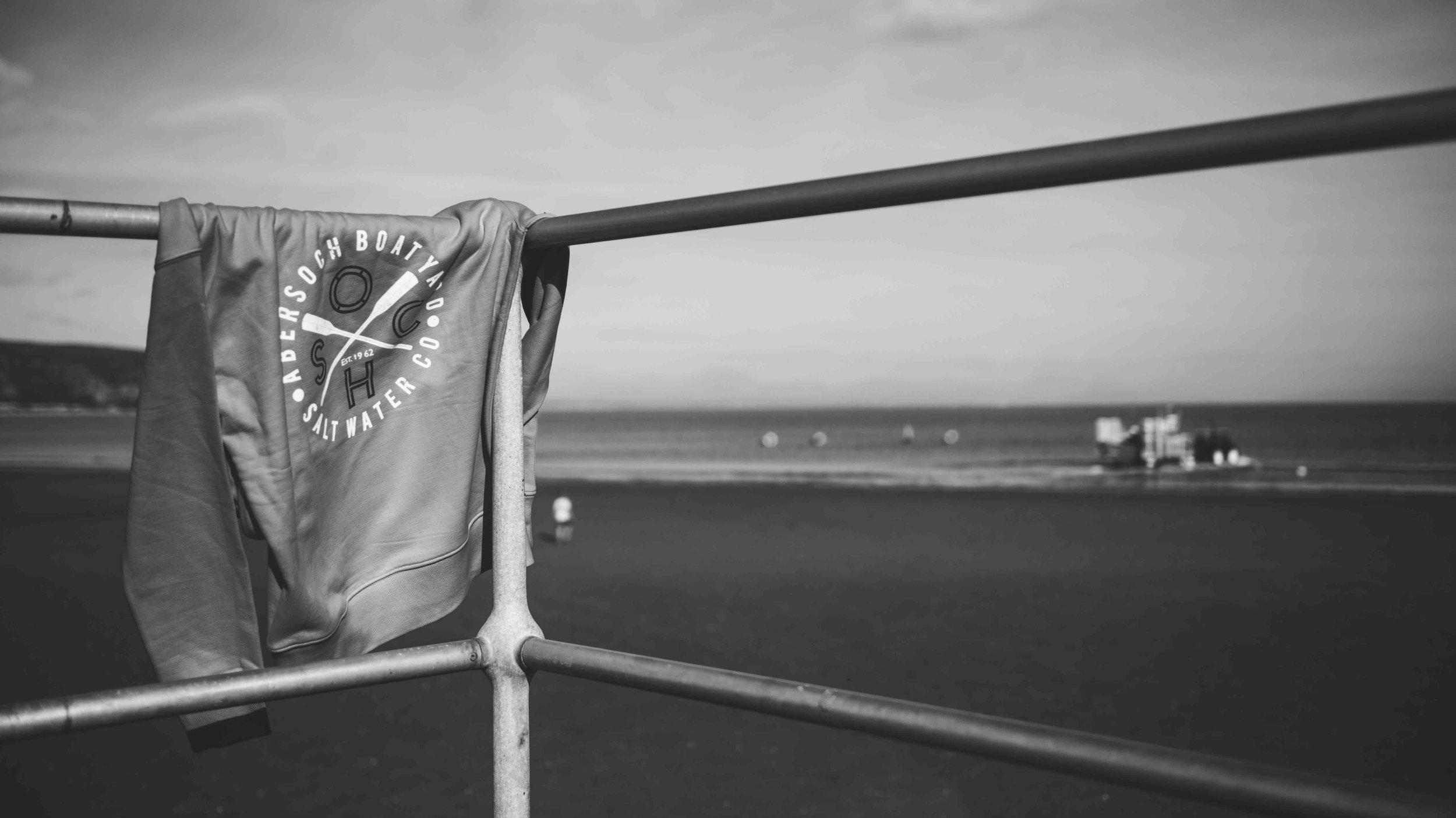 Monochrome photo of a sweatshirt draped over a metal railing with a beach and ocean in the background.