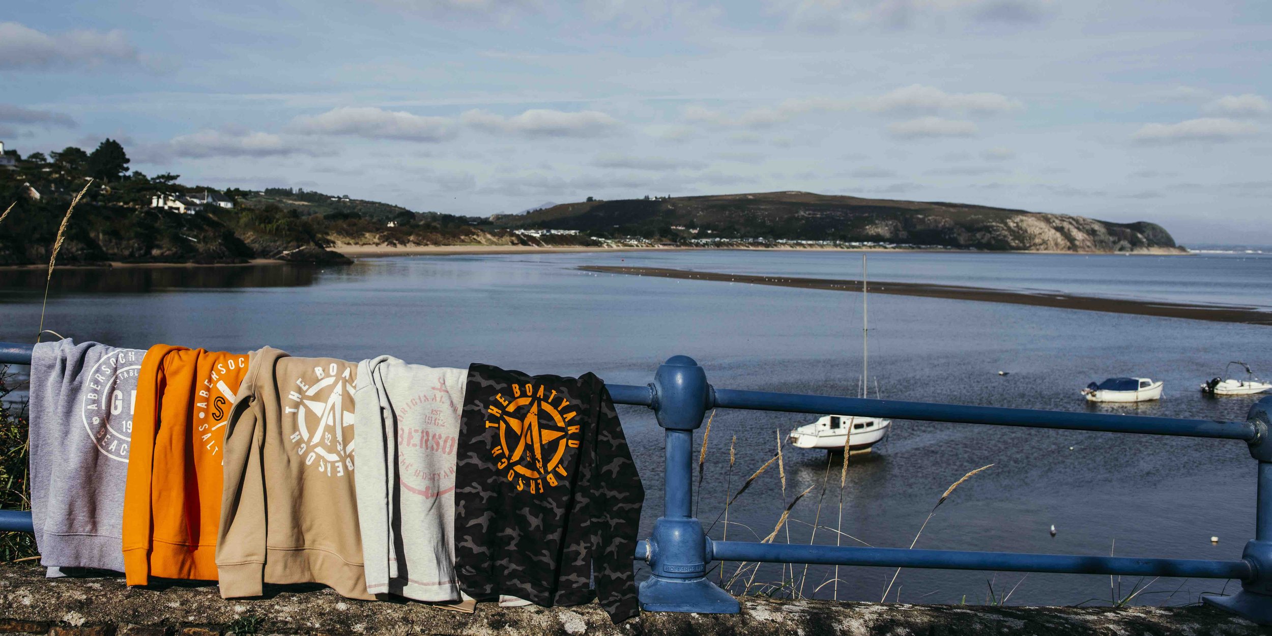 Five colorful sweatshirts hanging on a blue railing near a scenic coastal view with boats and hills in the background.