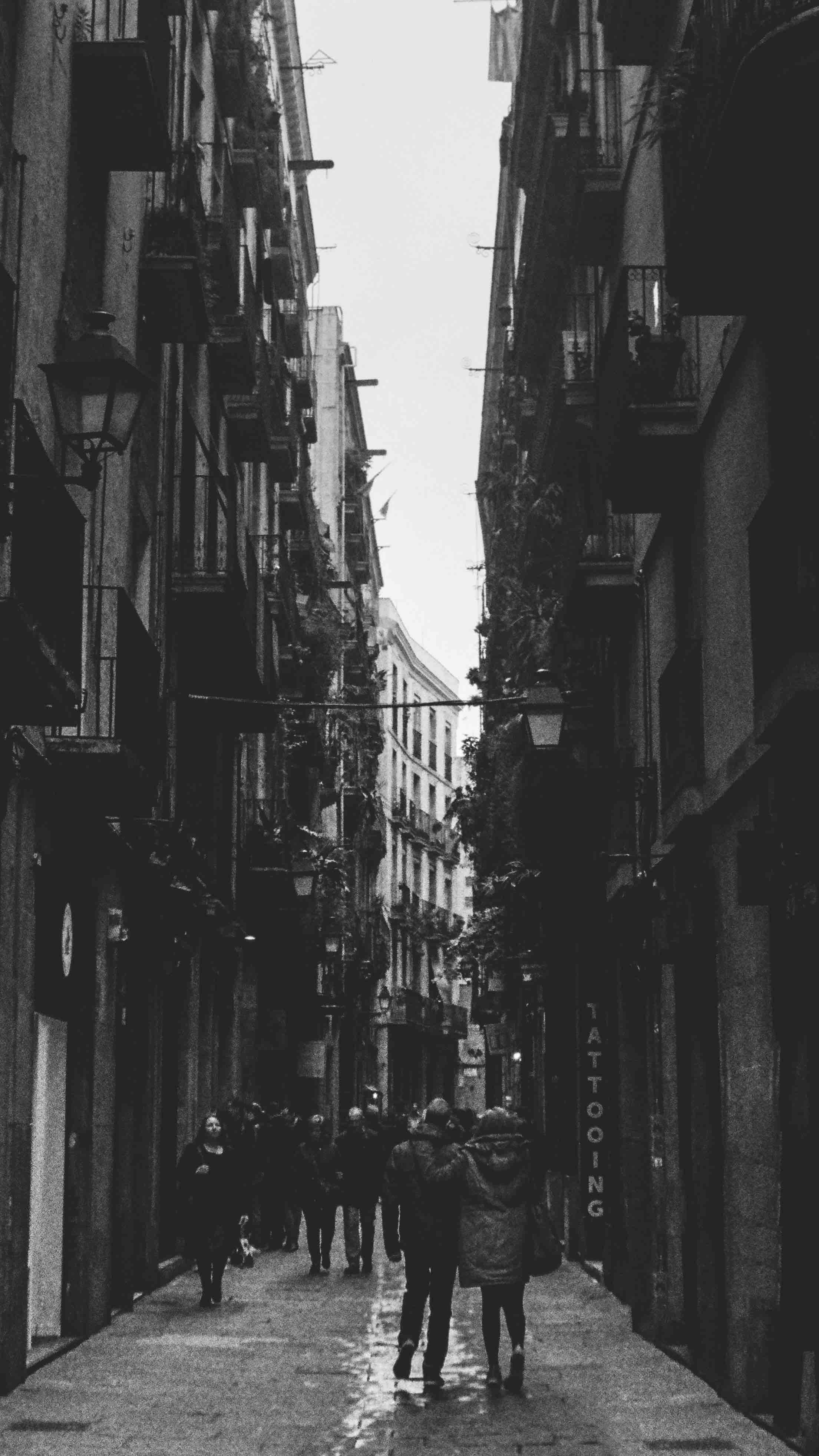 Black and white photo of a narrow city street lined with old buildings. People walking down the street, including a couple in the foreground. Some balconies have plants, and a tattoo parlor sign is visible on the right.