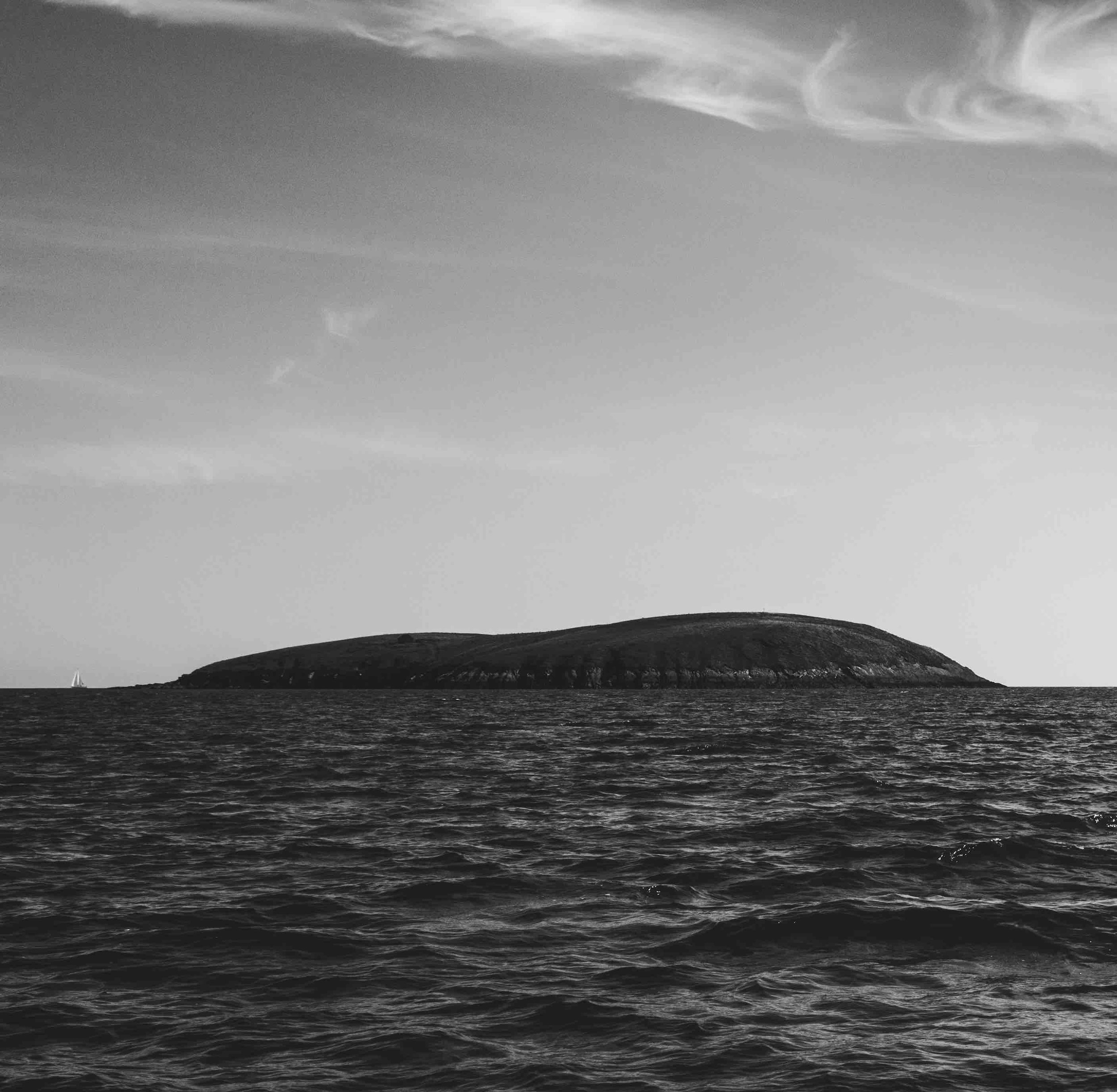 Black and white image of an island with rocky shoreline and calm ocean under a cloudy sky.