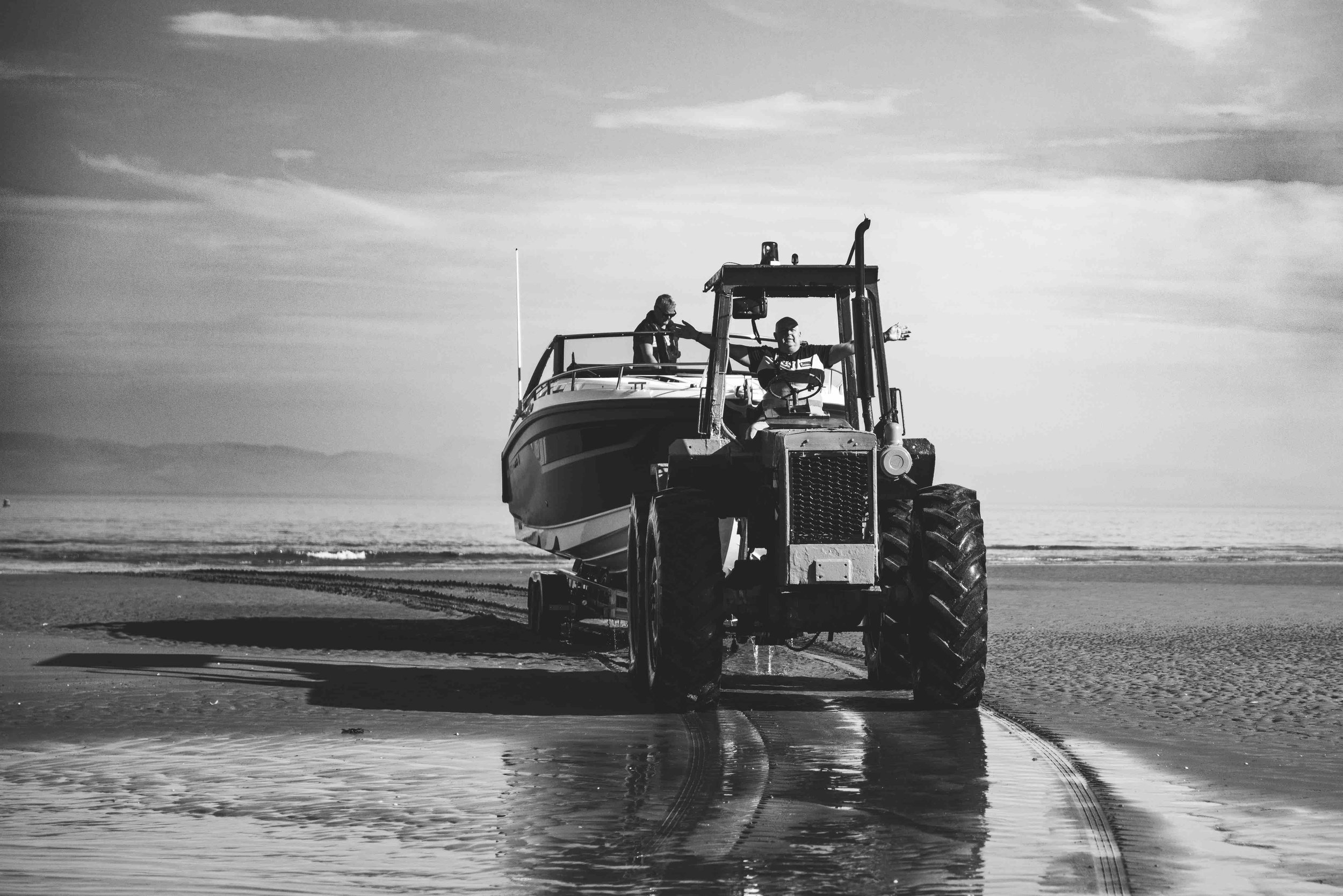 Black and white image of a tractor towing a boat on a beach with water and sky in the background.