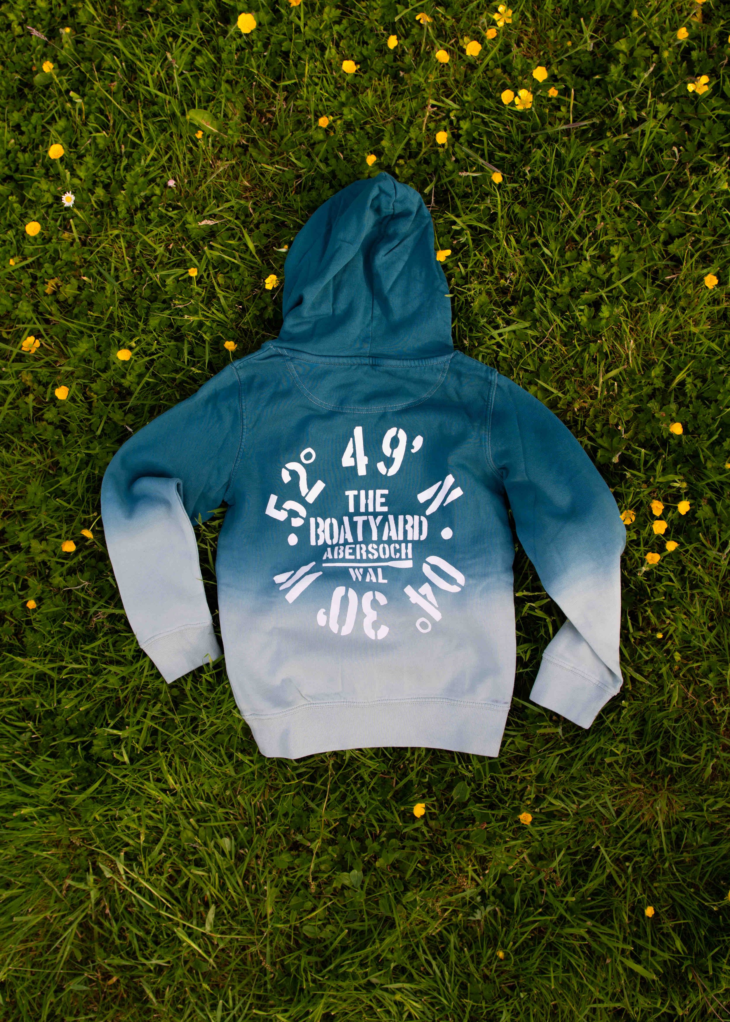 Blue hoodie with text and symbols, lying on grass with yellow flowers