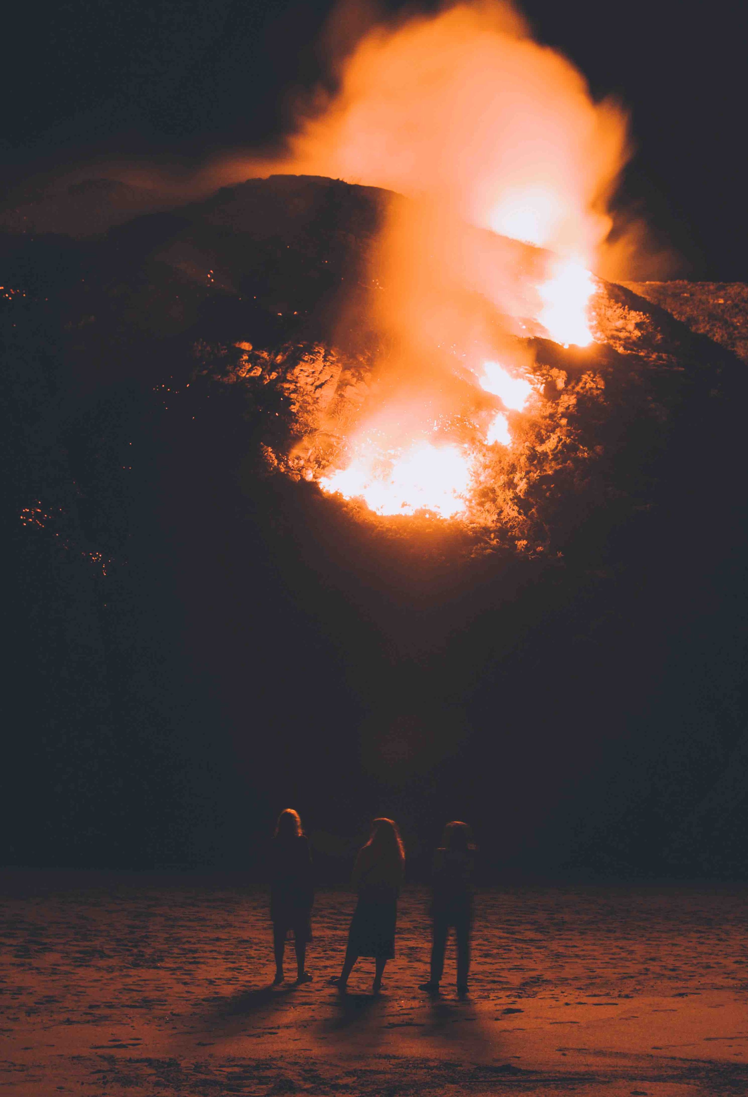 Night scene of three silhouetted people standing on a sand dune, watching a large fire on the hillside.